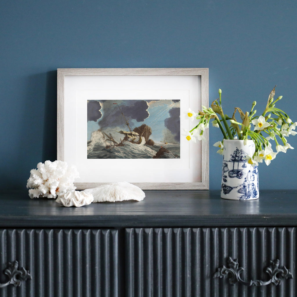 A framed vintage maritime painting of a stormy shipwreck scene is displayed on a dark wooden sideboard against a deep blue wall. The artwork, encased in a pale wood frame, depicts a ship struggling against powerful waves with dark storm clouds overhead. Beside the frame, a ceramic vase with nautical illustrations holds fresh yellow and white flowers. Coral pieces and seashells are arranged on the sideboard, enhancing the coastal aesthetic.