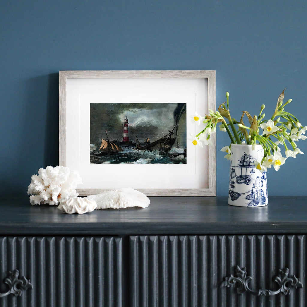 A framed nautical print featuring a stormy sea with a sinking ship near a red-and-white striped lighthouse. The artwork is displayed on a dark wooden sideboard, accompanied by white coral, a blue-and-white maritime-themed vase with fresh flowers, and a moody blue wall as the backdrop.