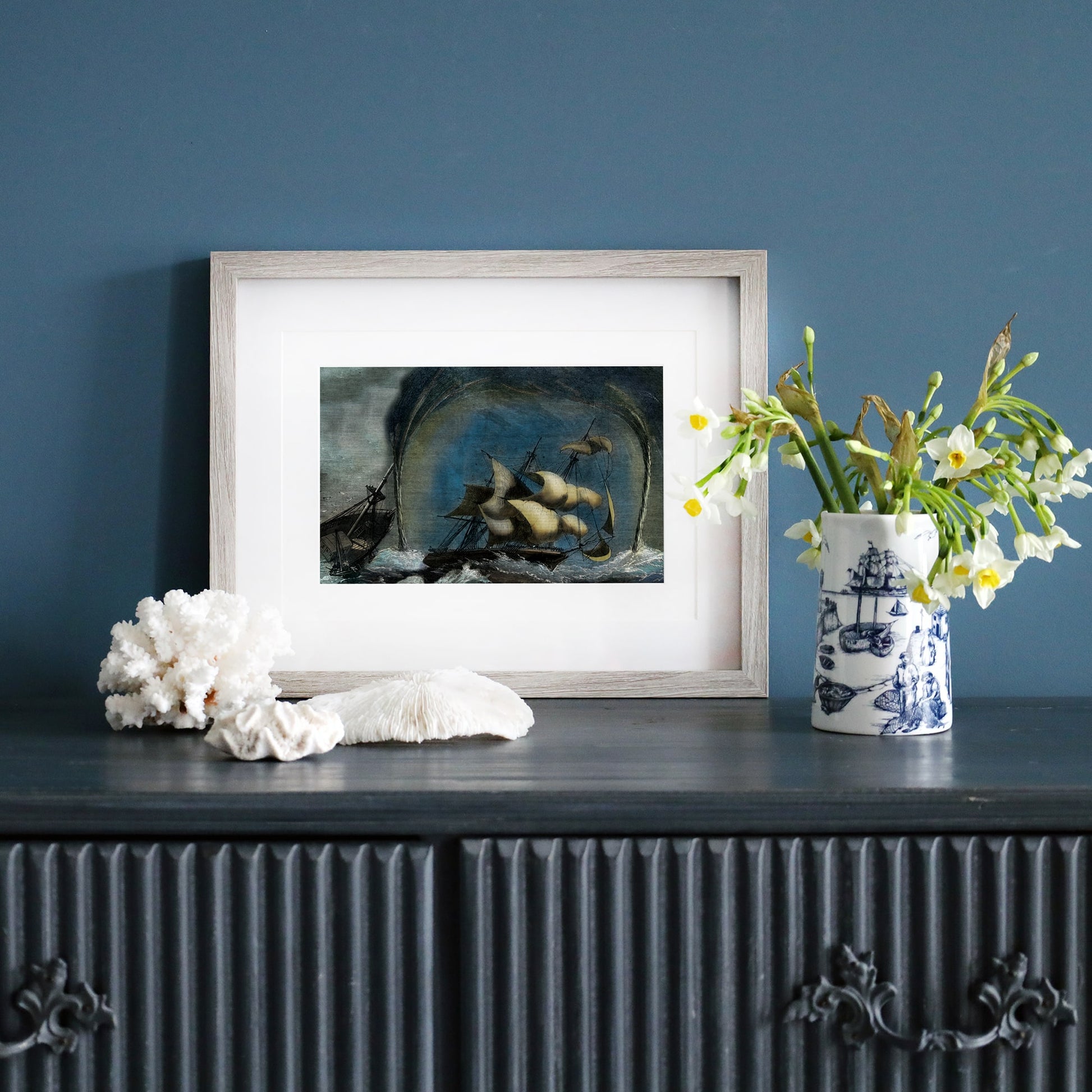 A framed maritime print featuring a dramatic ship caught in a storm with towering waterspouts, displayed on a dark wooden sideboard. The scene is complemented by nautical-themed decor, including a blue and white illustrated vase with fresh flowers, coral pieces, and a seashell, against a muted blue wall.