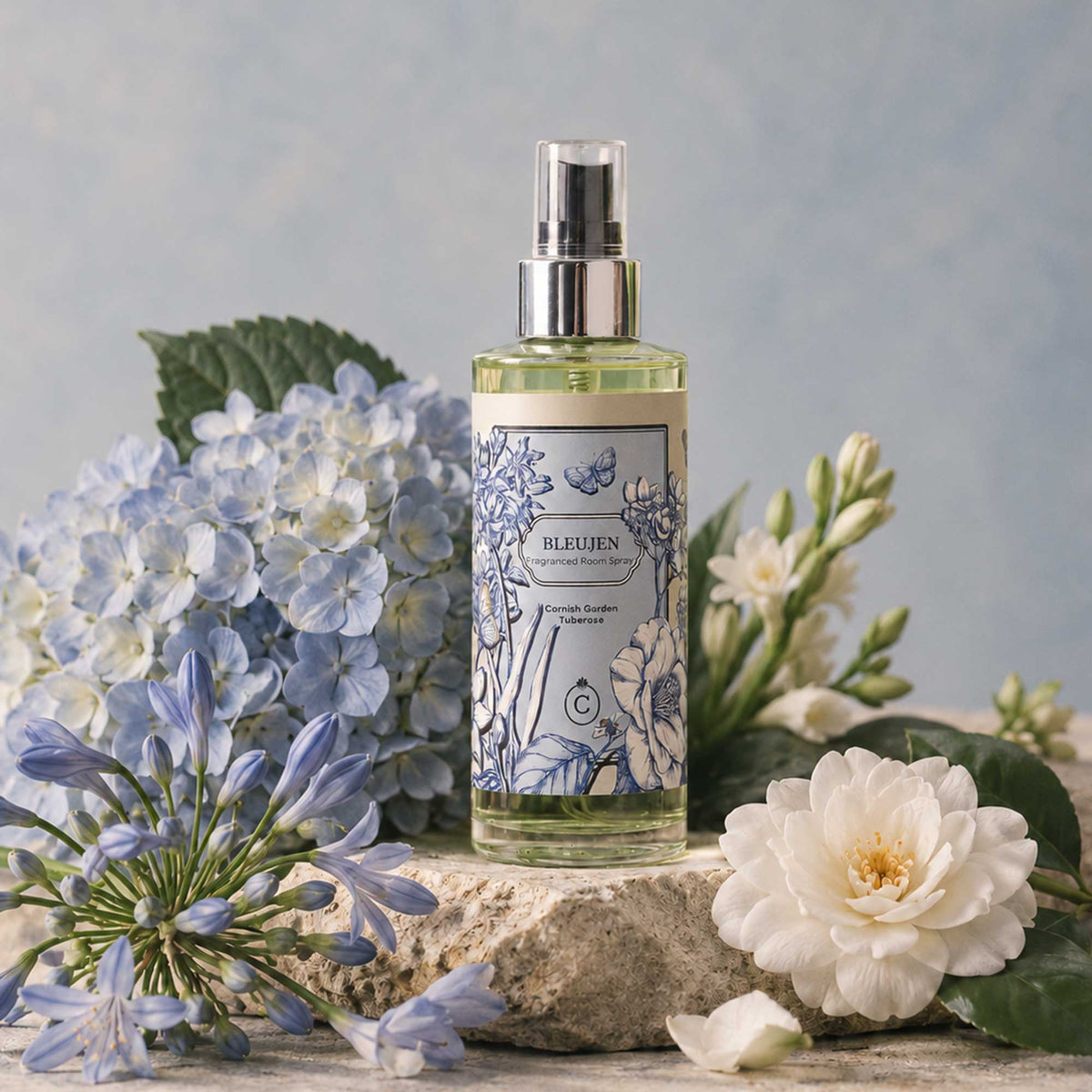 BLEUJEN Cornish Garden Tuberose fragranced room spray on natural stone, surrounded by soft blue hydrangeas, lavender-blue agapanthus, delicate white tuberose and a white camellia, against a muted blue-grey background.