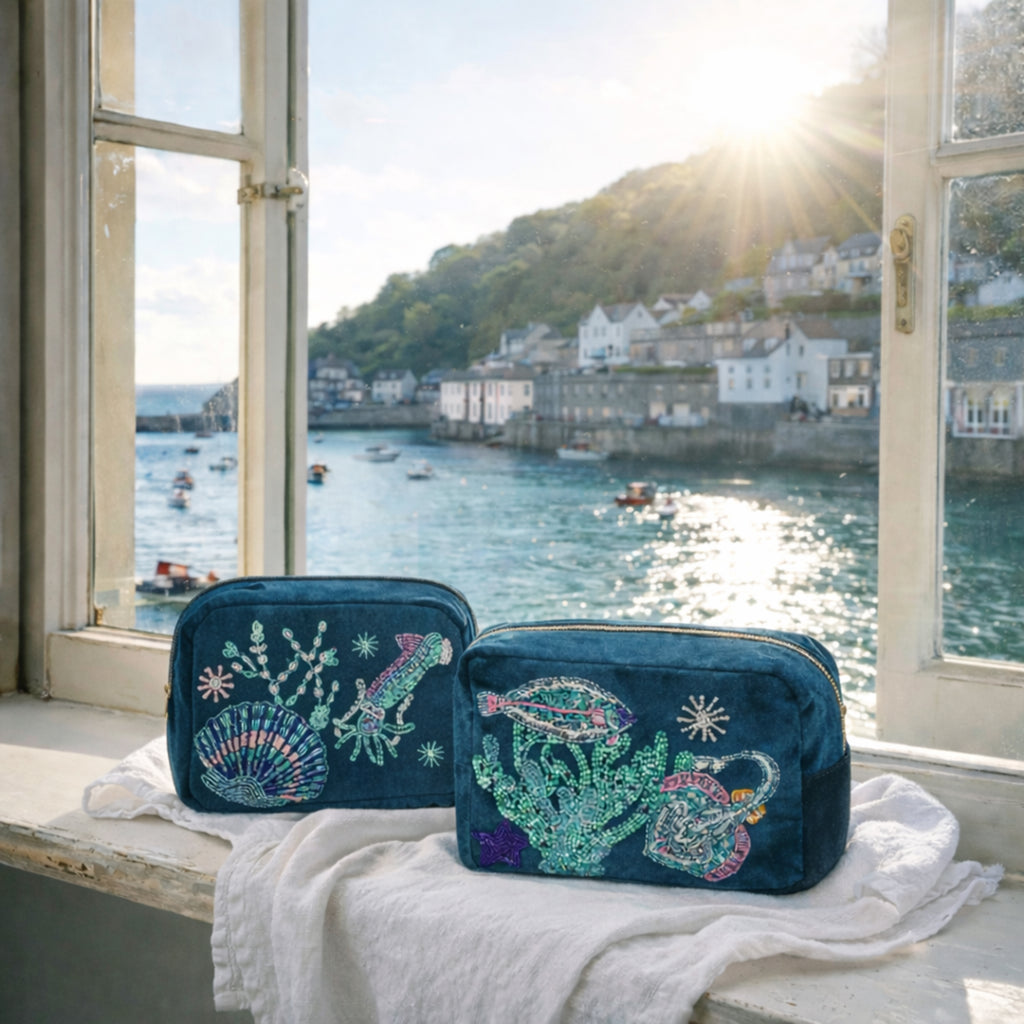 Two teal makeup bags with ocean-themed designs on a windowsill with a view of a harbour.