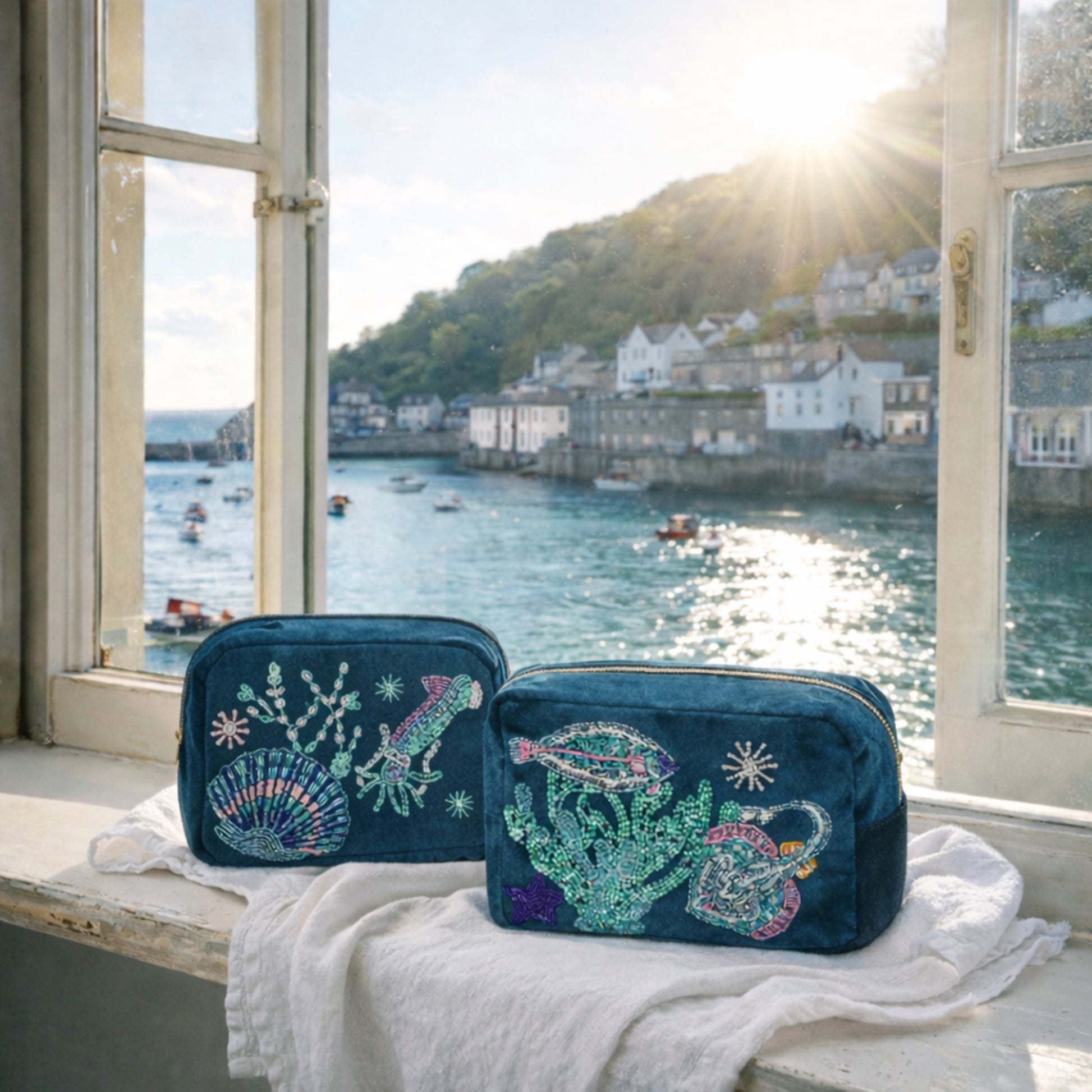 Two teal makeup bags with ocean-themed designs on a windowsill with a view of a harbour.