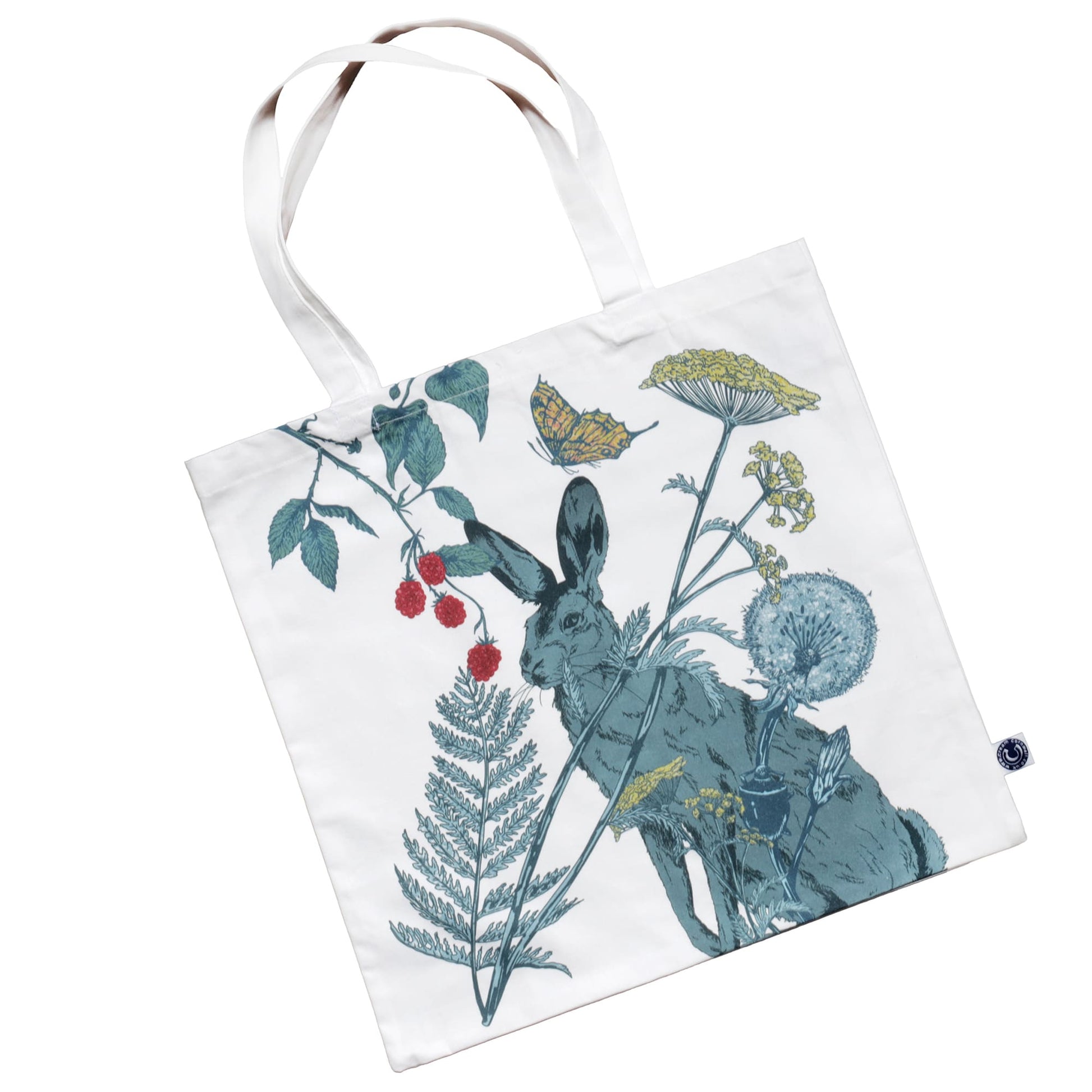 Fabric tote bag featuring a countryside-inspired illustration of a hare surrounded by wildflowers, ferns, dandelions, and raspberry vines. A butterfly adds a whimsical touch to the nature-themed design in shades of green, yellow, and red on a white background. The bag has sturdy white handles.