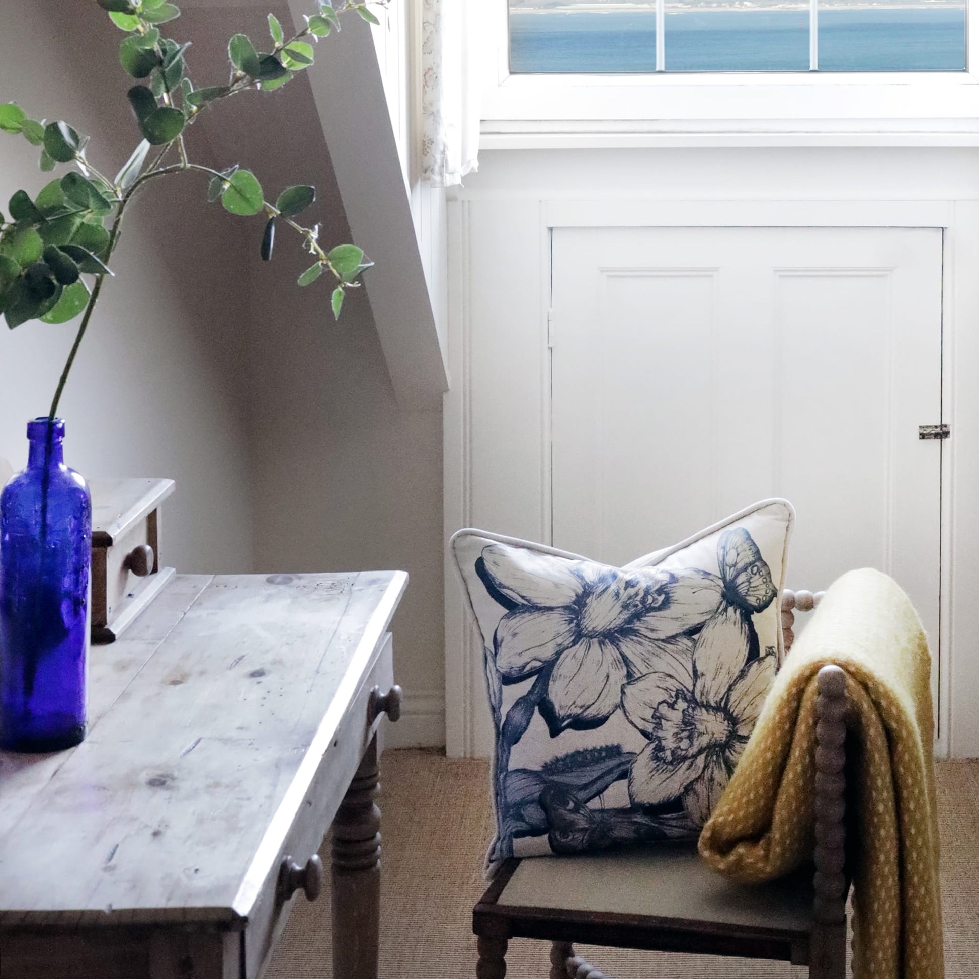 Chair with floral daffodil pillow and yellow blanket next to a wooden table with a blue bottle and plant.