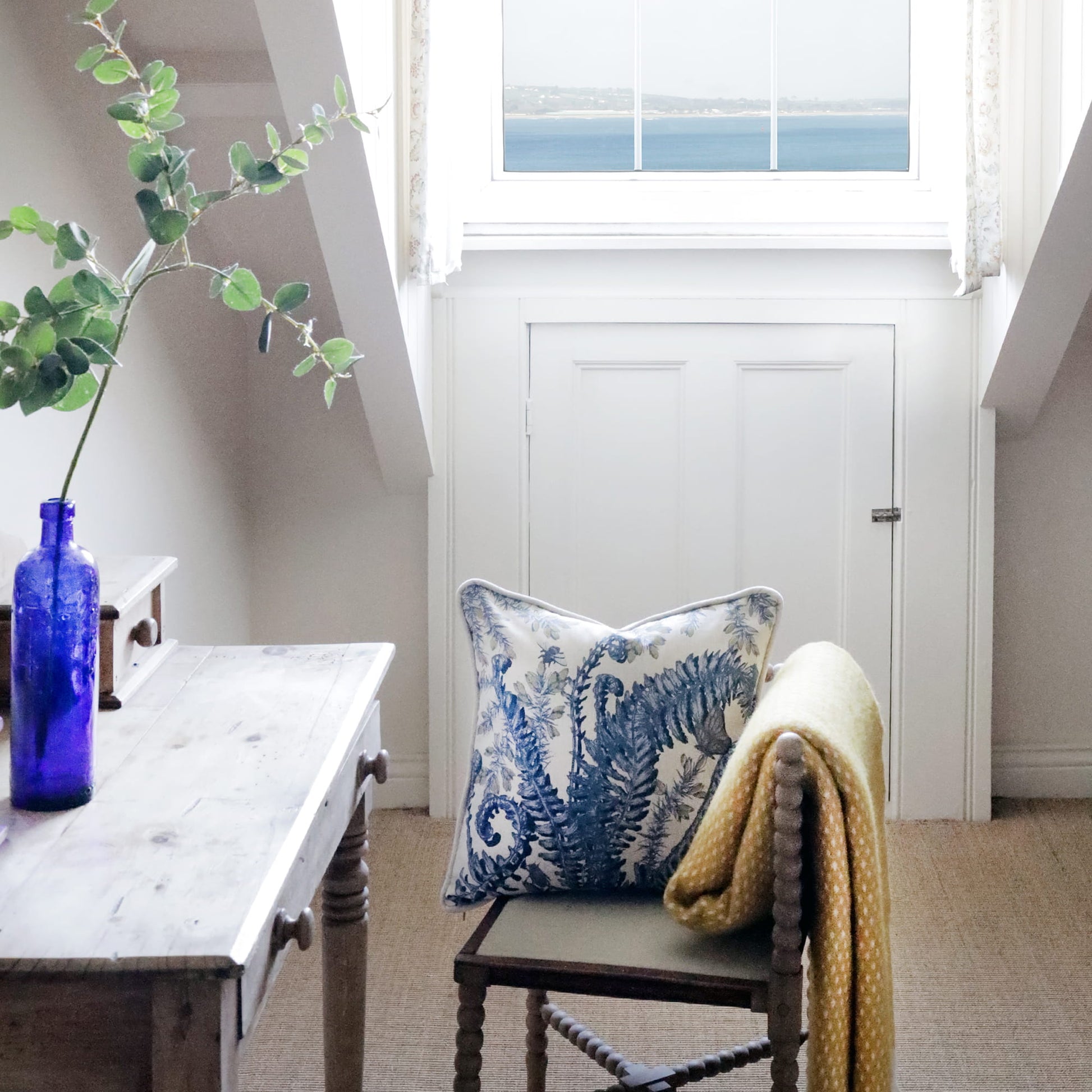Nook with a chair featuring a blue fern & gorse patterned pillow, a yellow blanket, and a blue vase on a table.