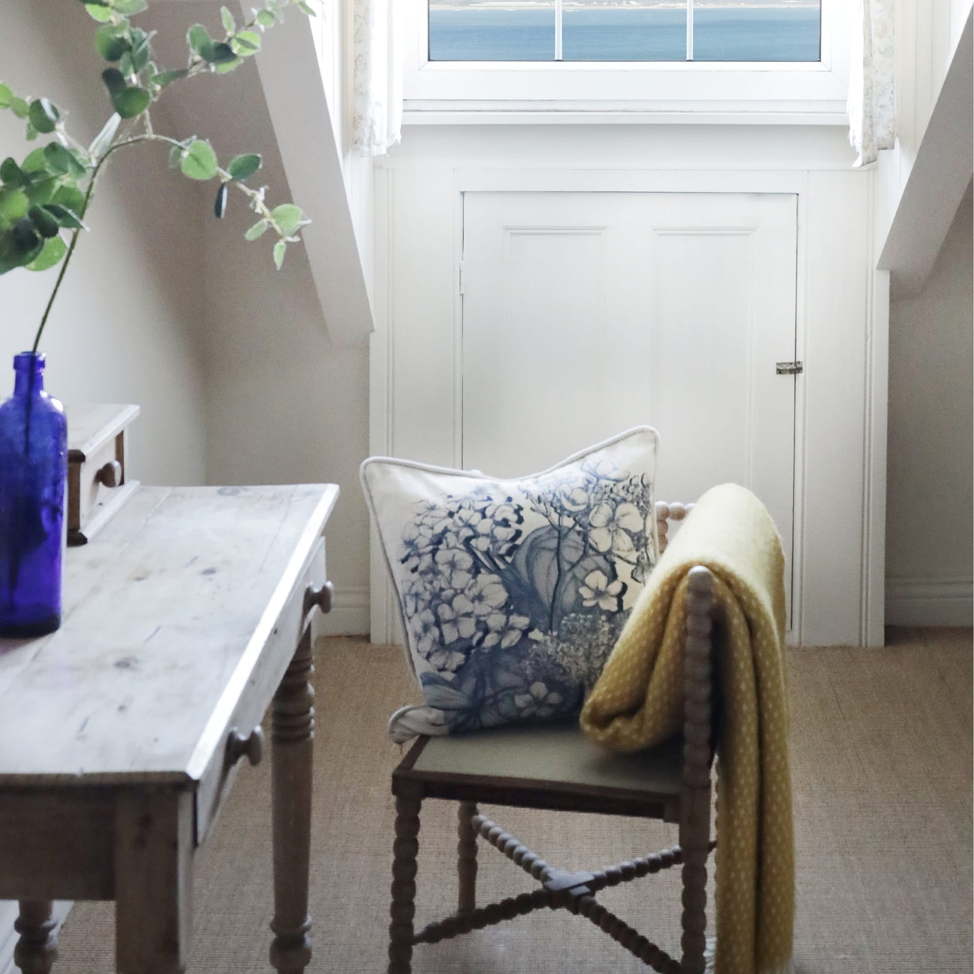 Chair with a decorative hydrangea pillow and yellow blanket in a room with a wooden table and window.