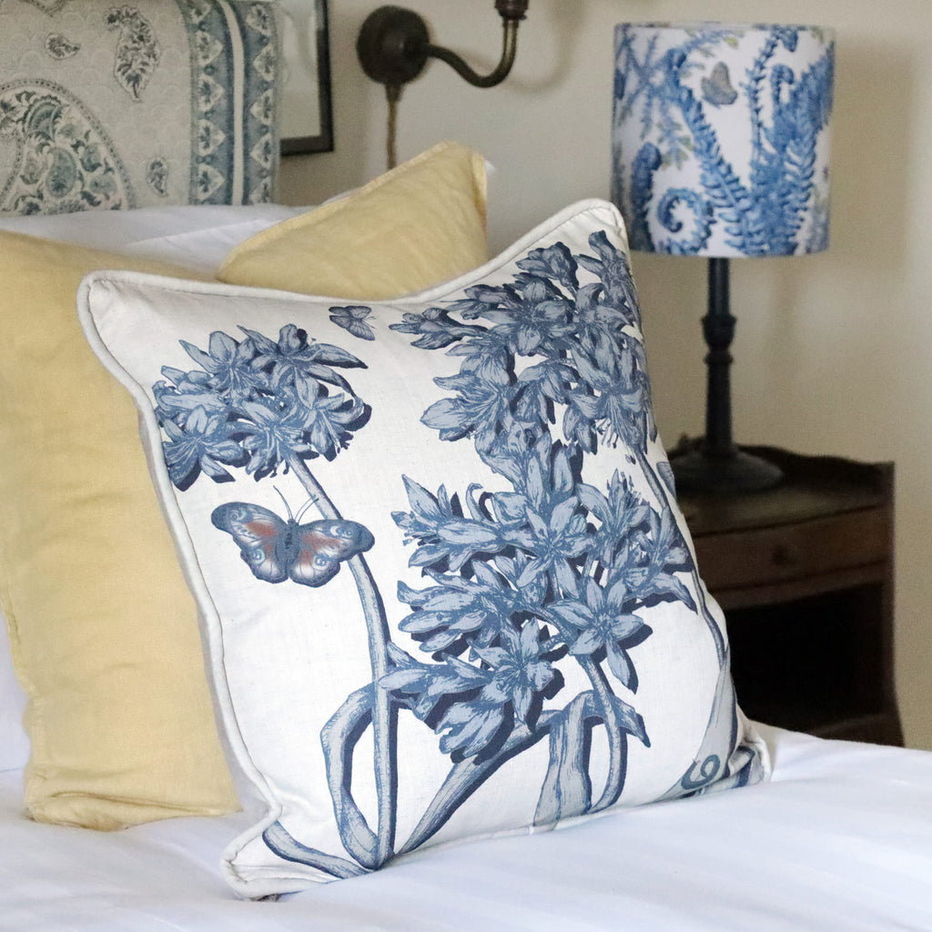 Decorative pillow with blue agapanthus floral and butterfly design on a bed with yellow and white pillows.