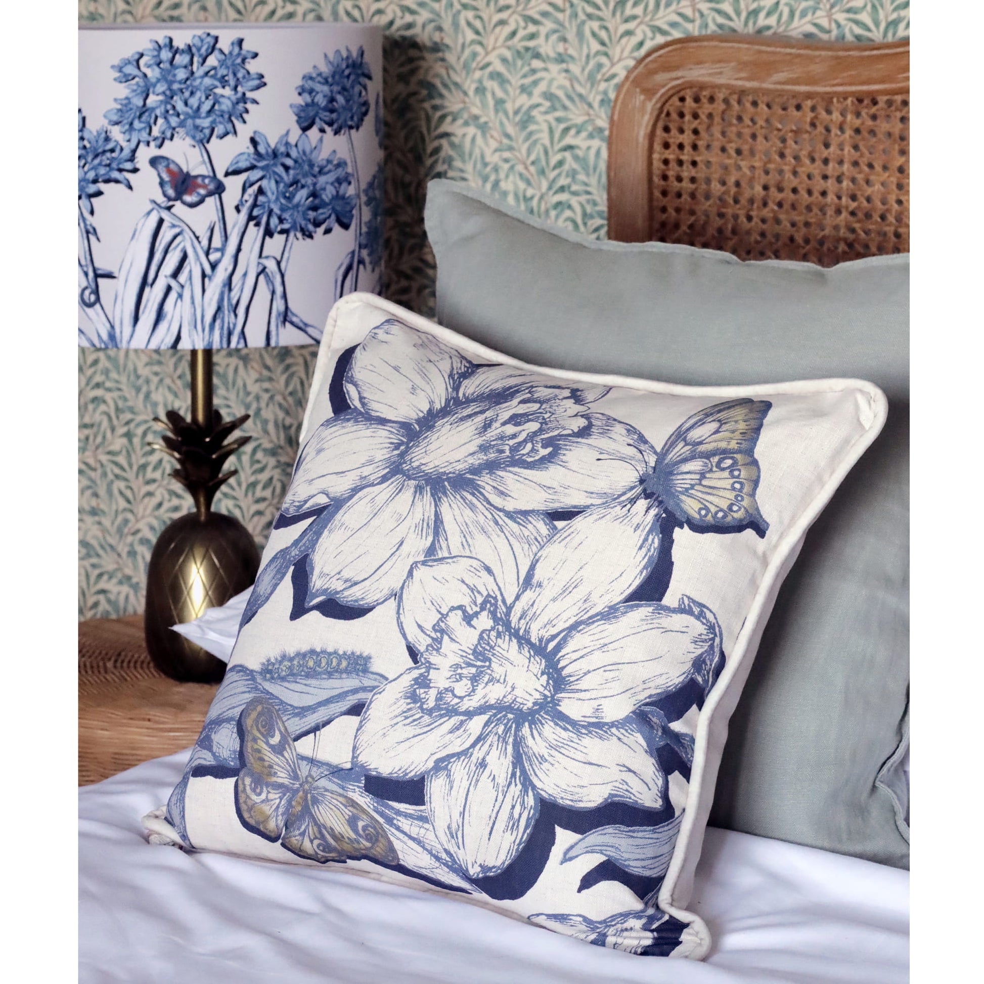 Decorative pillow with blue floral and butterfly design on a bed with floral wallpaper.
