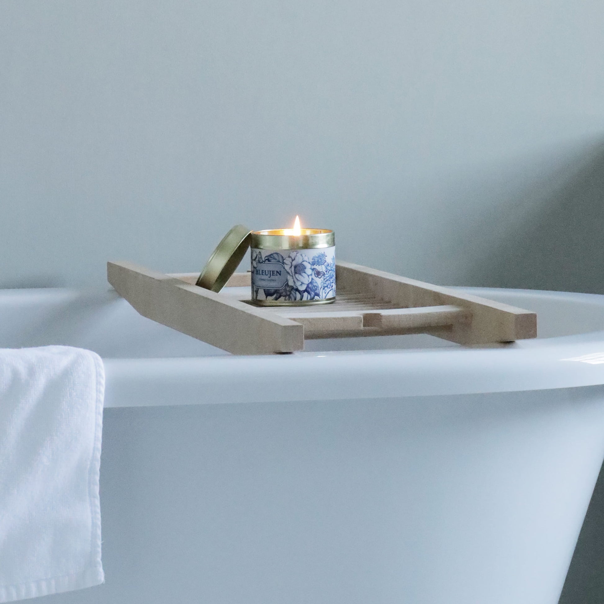 Bleujen candle in a decorative tin on a wooden tray over a bathtub with a towel.