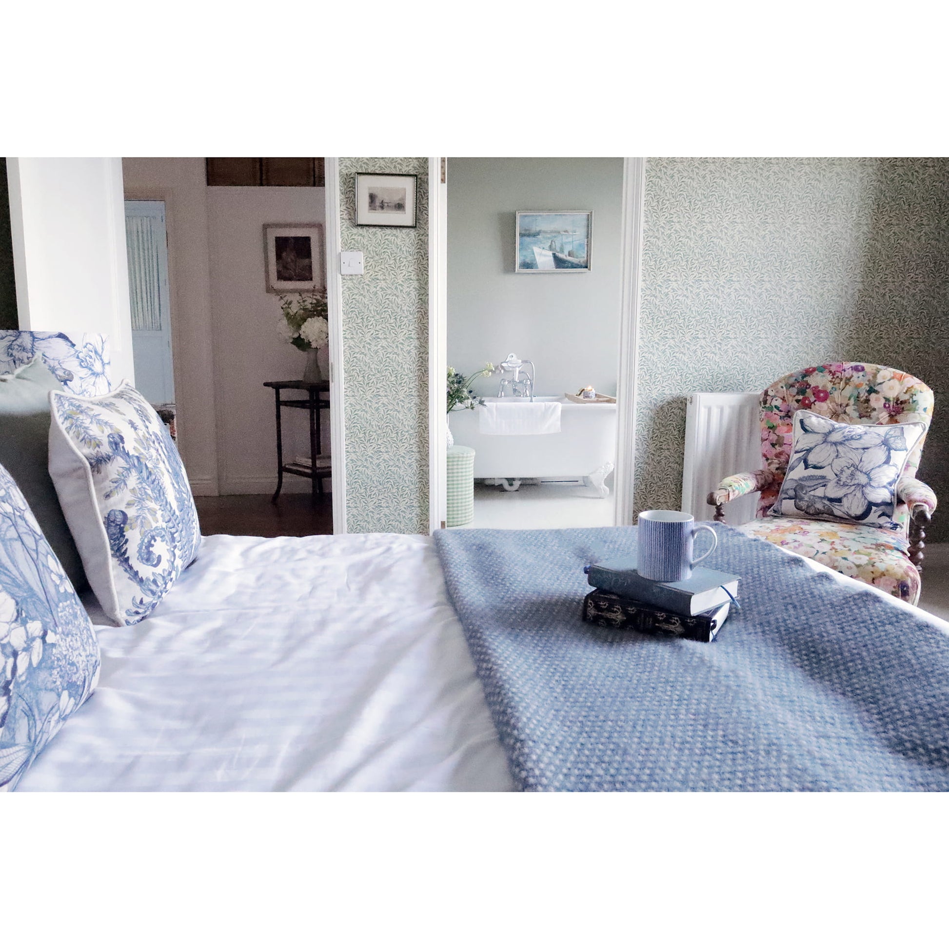 Bed with blue and white bedding, floral armchair, and bathroom in the background.