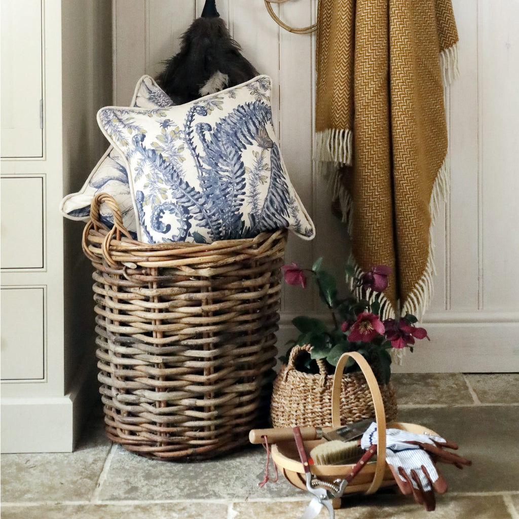Wicker laundry basket with decorative fern & gorse cushions and a plant in a home setting.