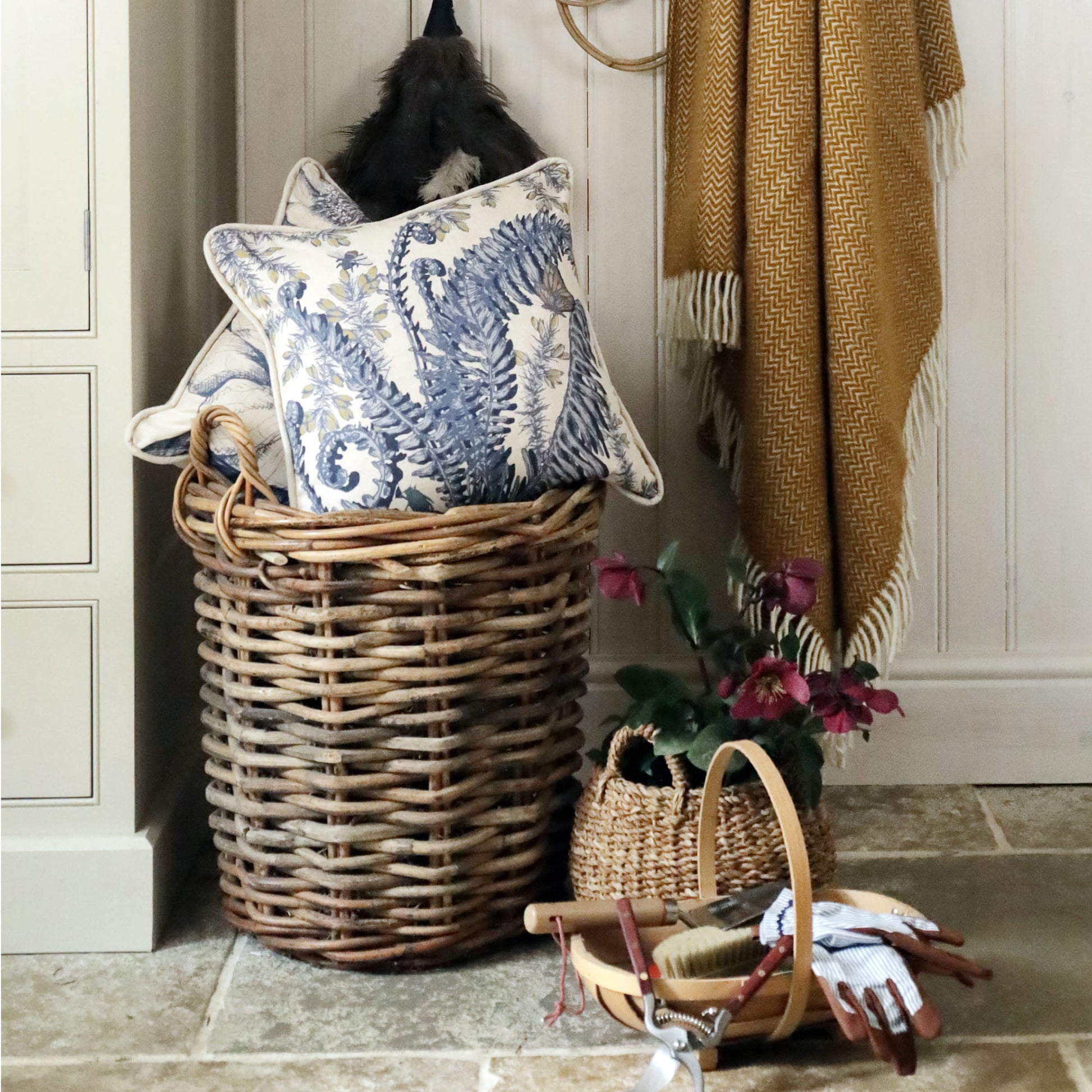 Wicker laundry basket with decorative fern & gorse cushions and a plant in a home setting.
