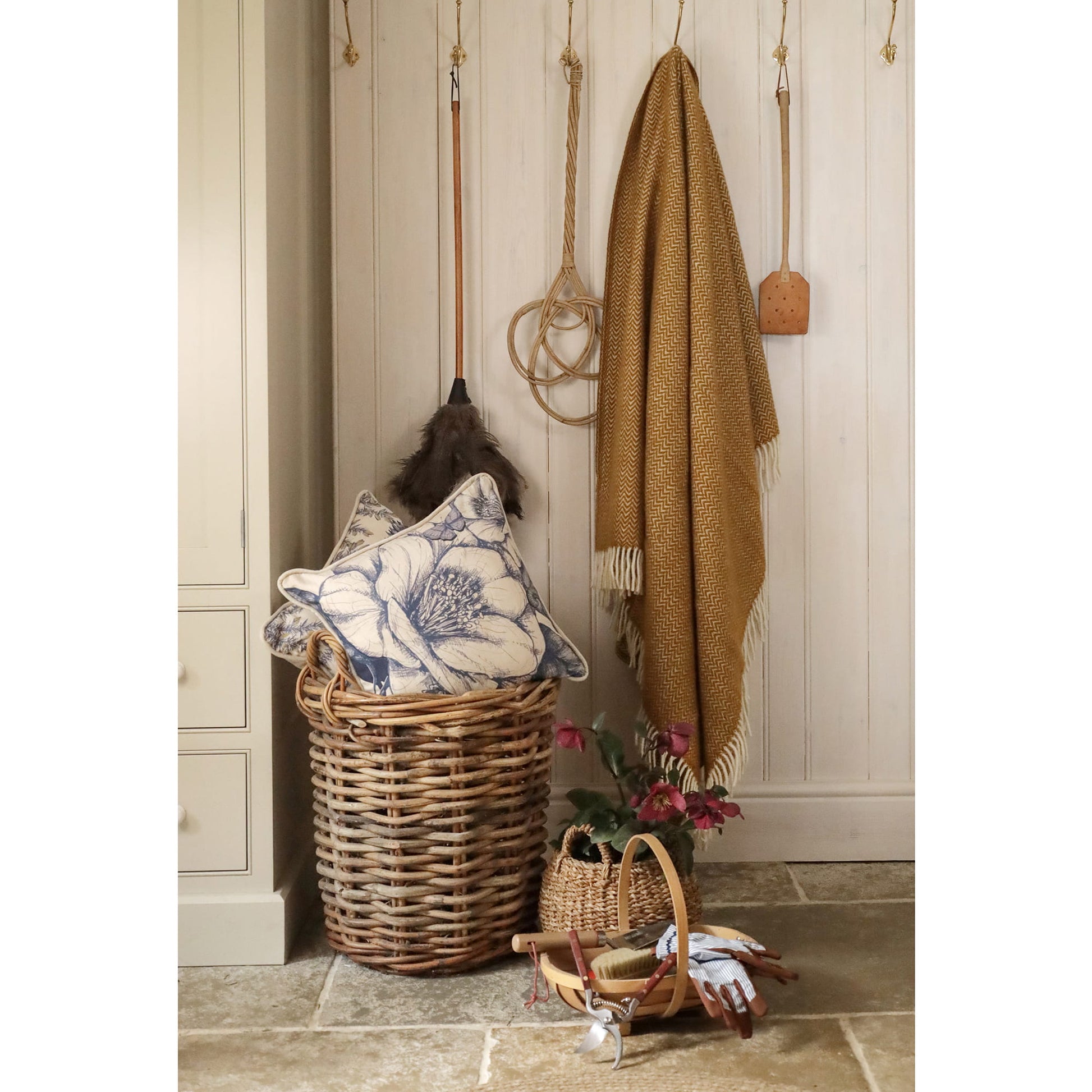 Wicker basket with decorative pillow, blanket, and small plant against a wooden wall.