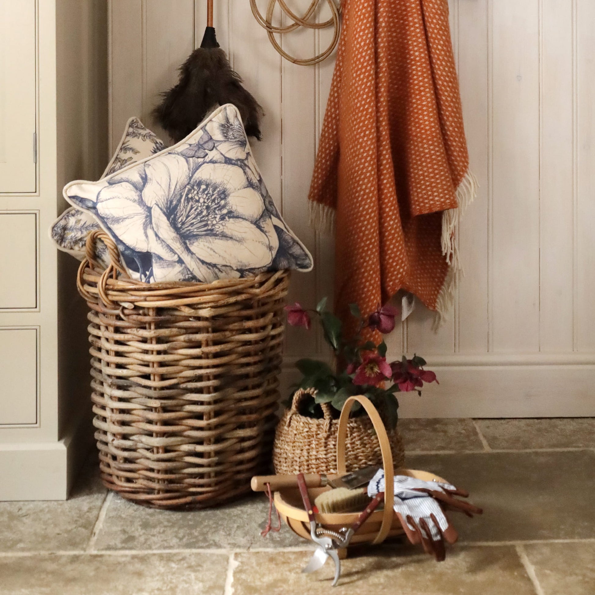 Wicker laundry basket with camelia floral-patterned pillow, orange blanket, and small basket on tiled floor.