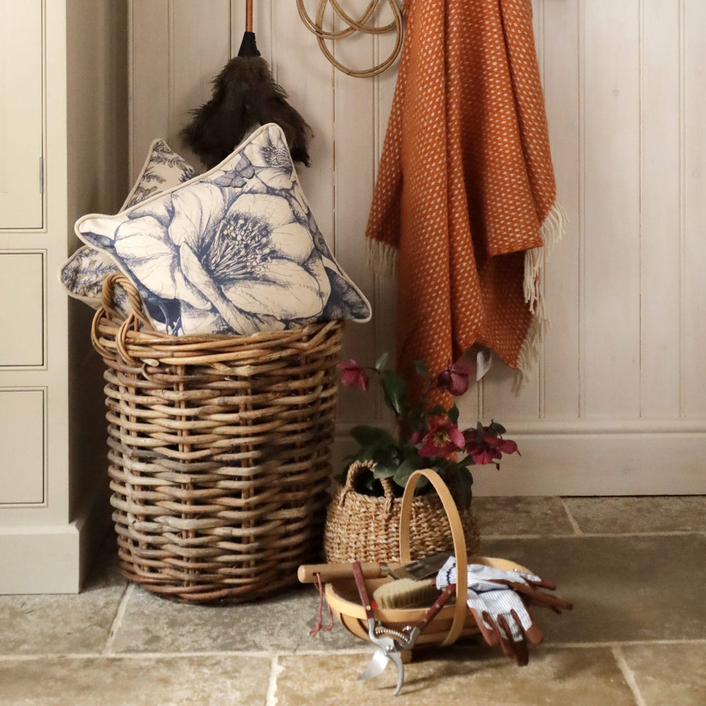 Wicker laundry basket with floral-patterned pillow, orange blanket, and small basket on tiled floor.