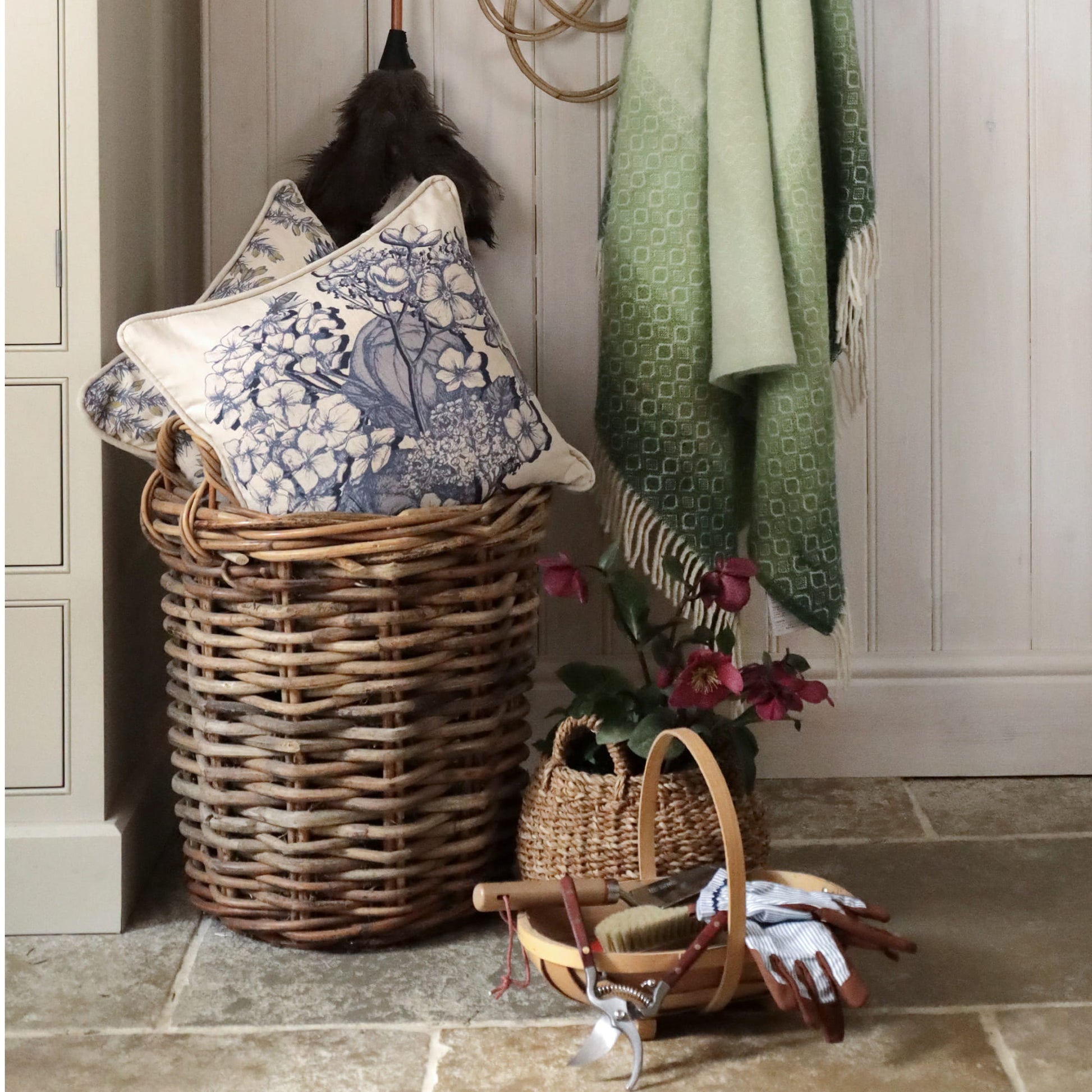 Wicker laundry basket with decorative pillows and a green towel on a wooden floor.