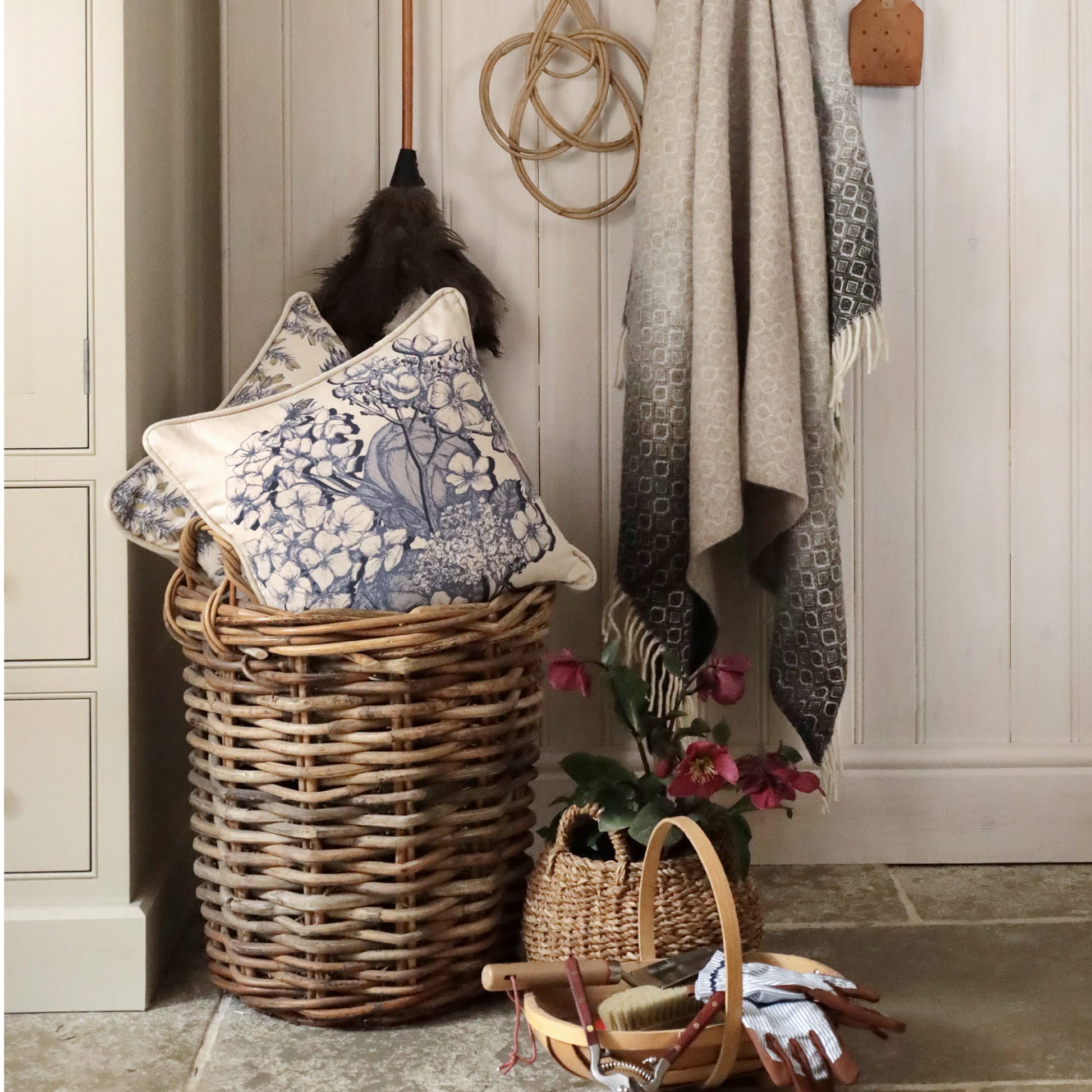 Wicker laundry basket with decorative pillows, plant, and folded clothes against a wooden wall.