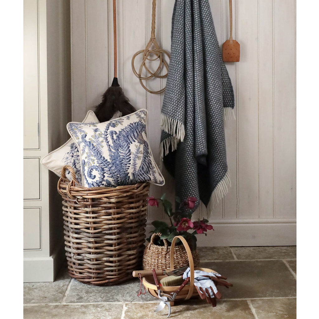Wicker basket with decorative pillows and a blanket against a wooden wall.