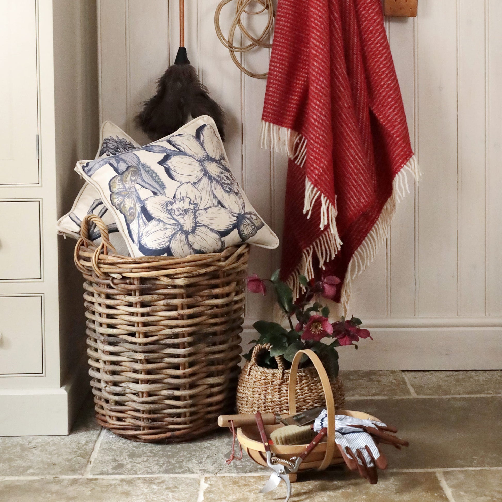 Wicker laundry basket with floral pillows, red blanket, and gardening tools in a home setting.