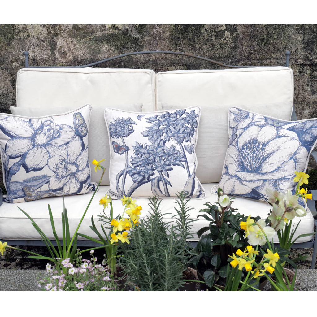 White outdoor sofa with blue agapanthus ,daffodil & camelia floral cushions surrounded by plants and flowers. 