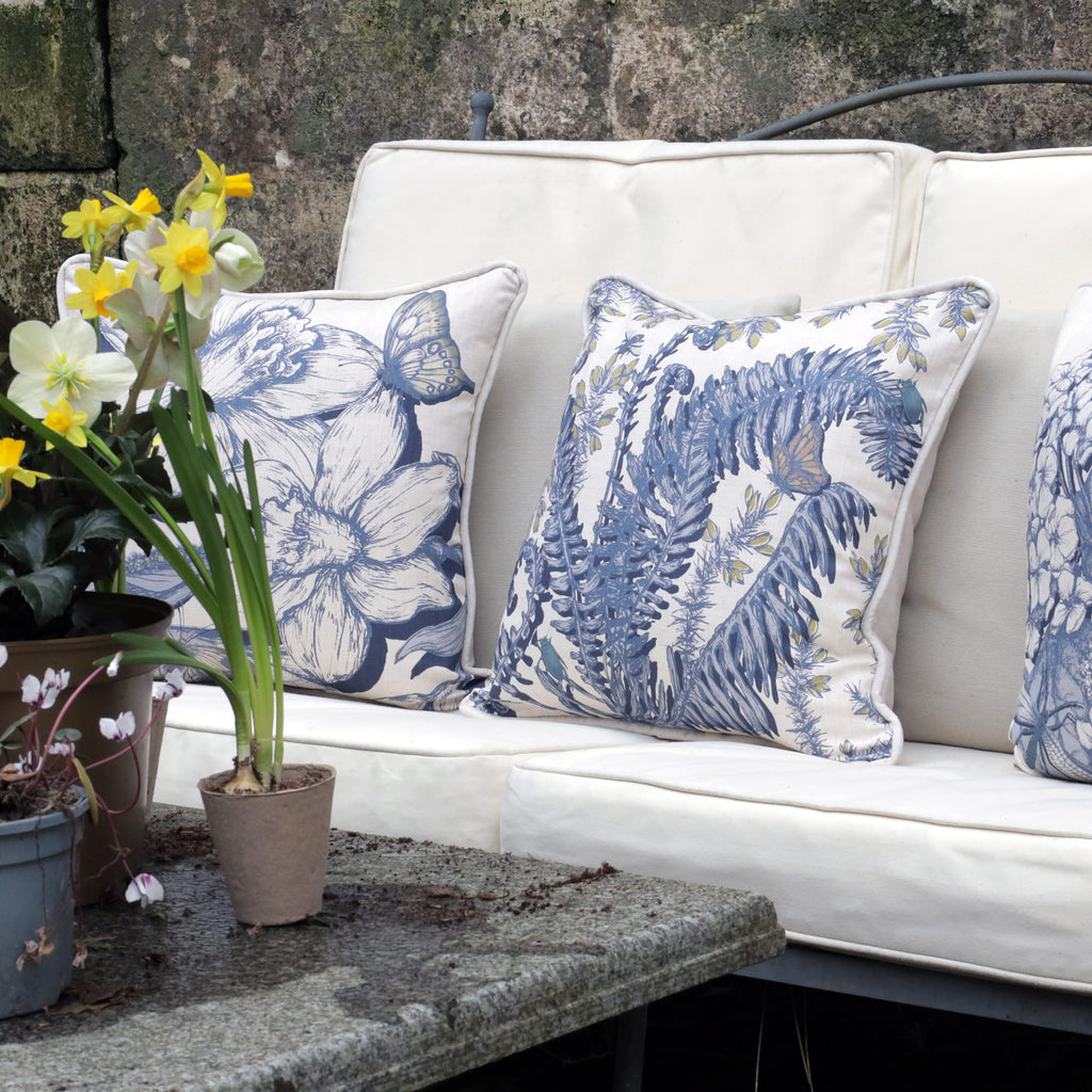 White outdoor sofa with blue fern & daffodil floral patterned cushions, surrounded by potted plants.