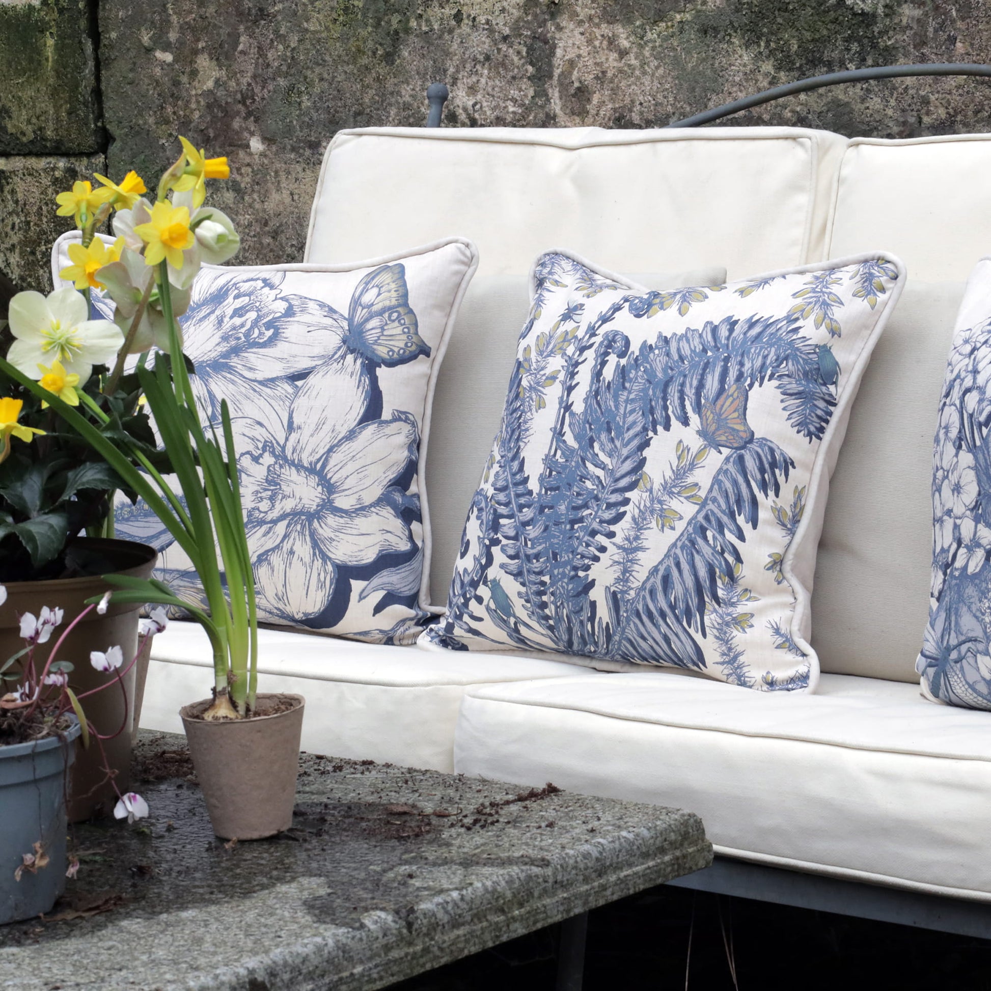White outdoor sofa with blue fern & daffodil floral patterned cushions, surrounded by potted plants.