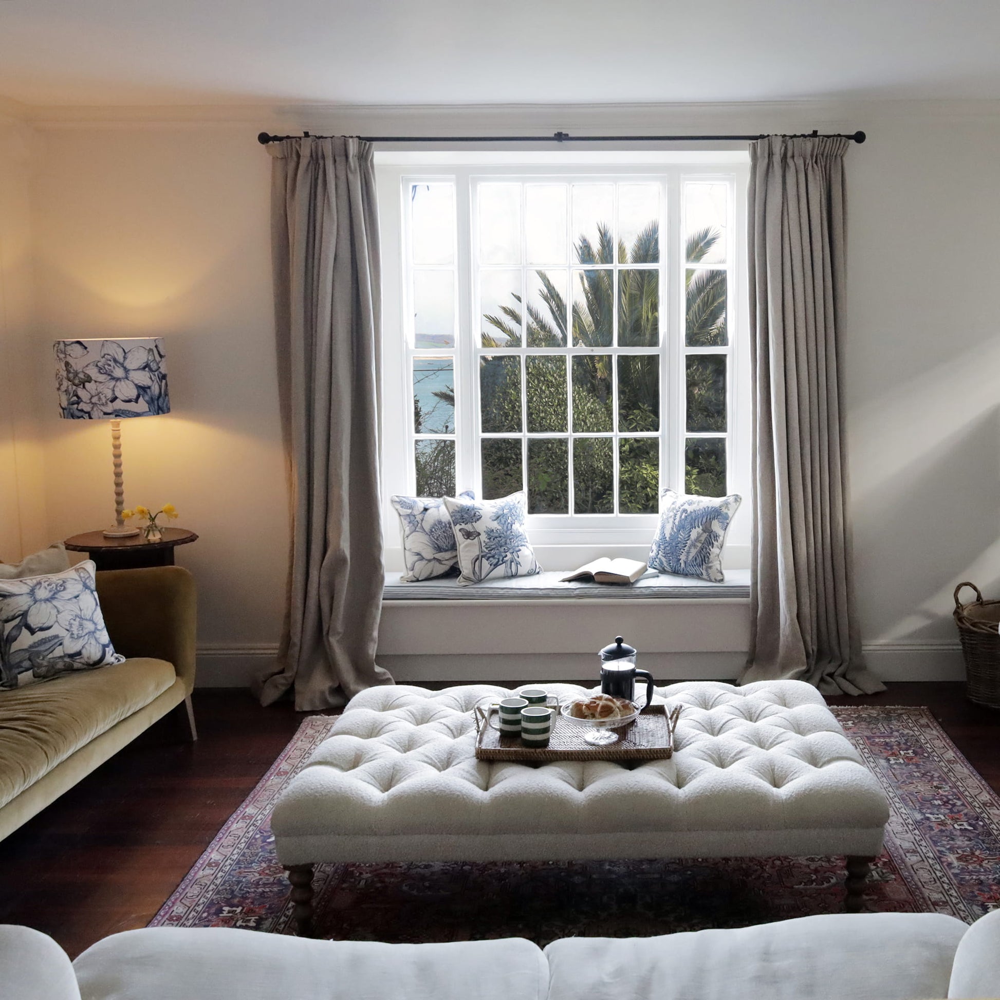Living room with a white ottoman, sofa, and cornish  floral cuahions on window seat with curtains.
