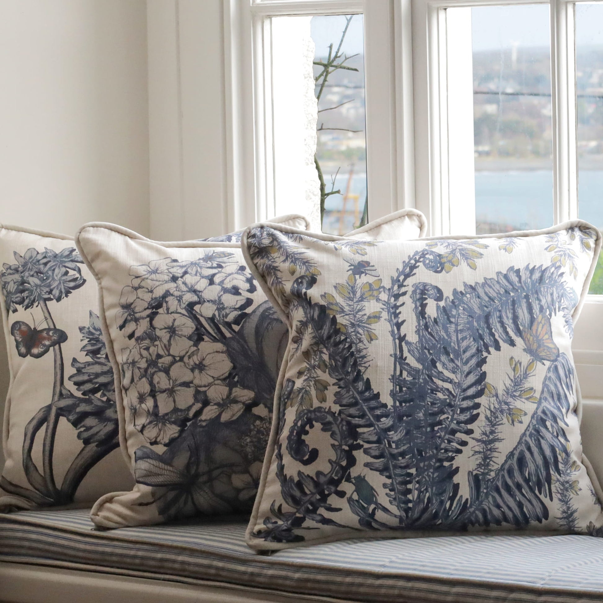 Decorative pillows with fern, hydrangea & agapanthus floral patterns on a window seat with a view of water.