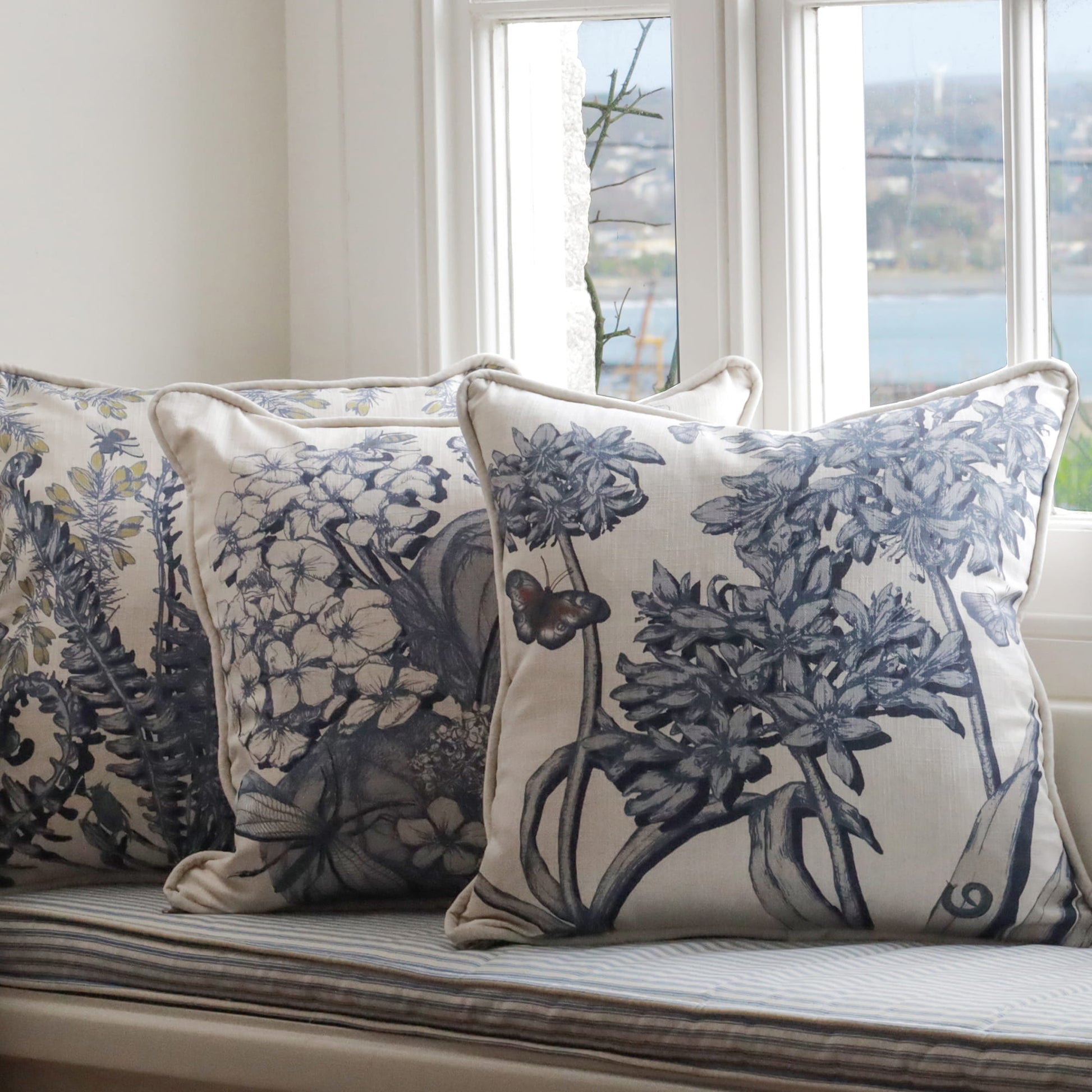 Decorative pillows with agapanthus, hydrangea & fern floral patterns on a window seat with a view of water.
