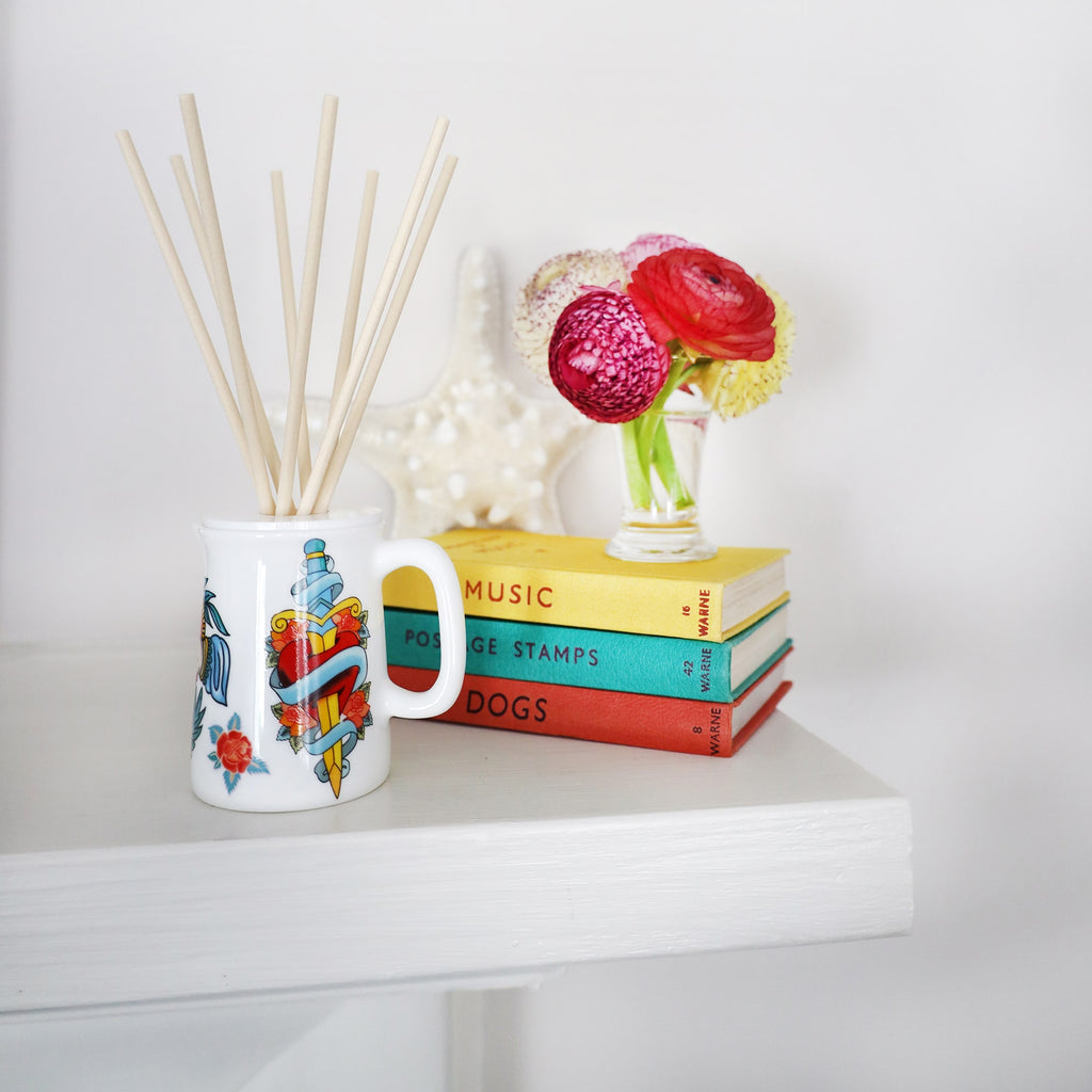 Sailors Story room diffuser with natural reeds on a shelf, in front of a set of three books and a small vase of flowers.