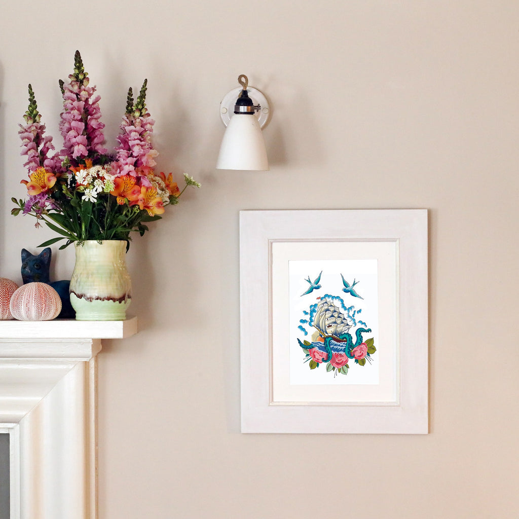 A framed nautical print hangs on a light-colored wall, featuring a tattoo-style illustration of a sailing ship caught in the tentacles of a kraken, with swallows and roses surrounding it. The frame is positioned above a mantel decorated with a vase of colorful flowers, a ceramic cat, and textured decorative spheres. A wall-mounted light fixture is above the artwork.