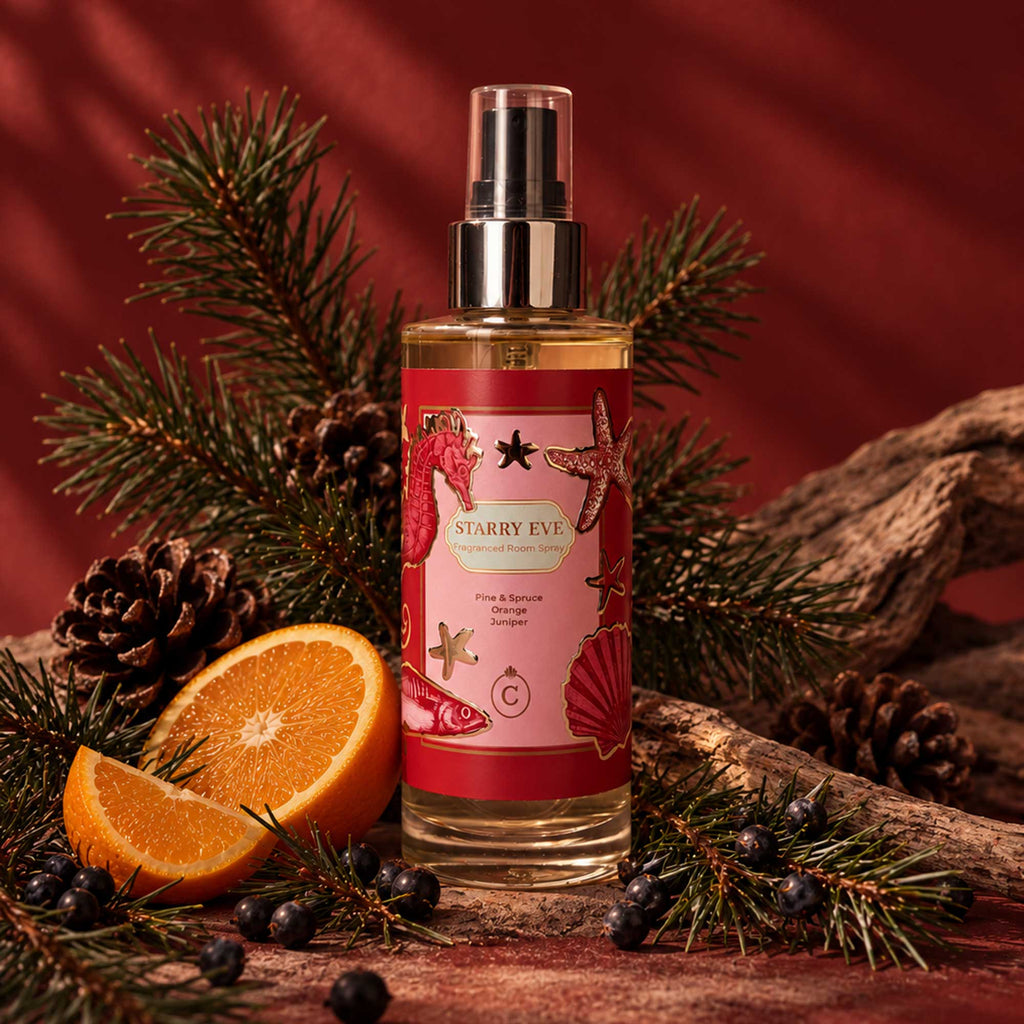 Starry Eve fragranced room spray in a red and pink bottle with gold detailing, surrounded by fresh pine and spruce branches, pinecones, juniper berries, orange slices and rustic wood against a warm red background.