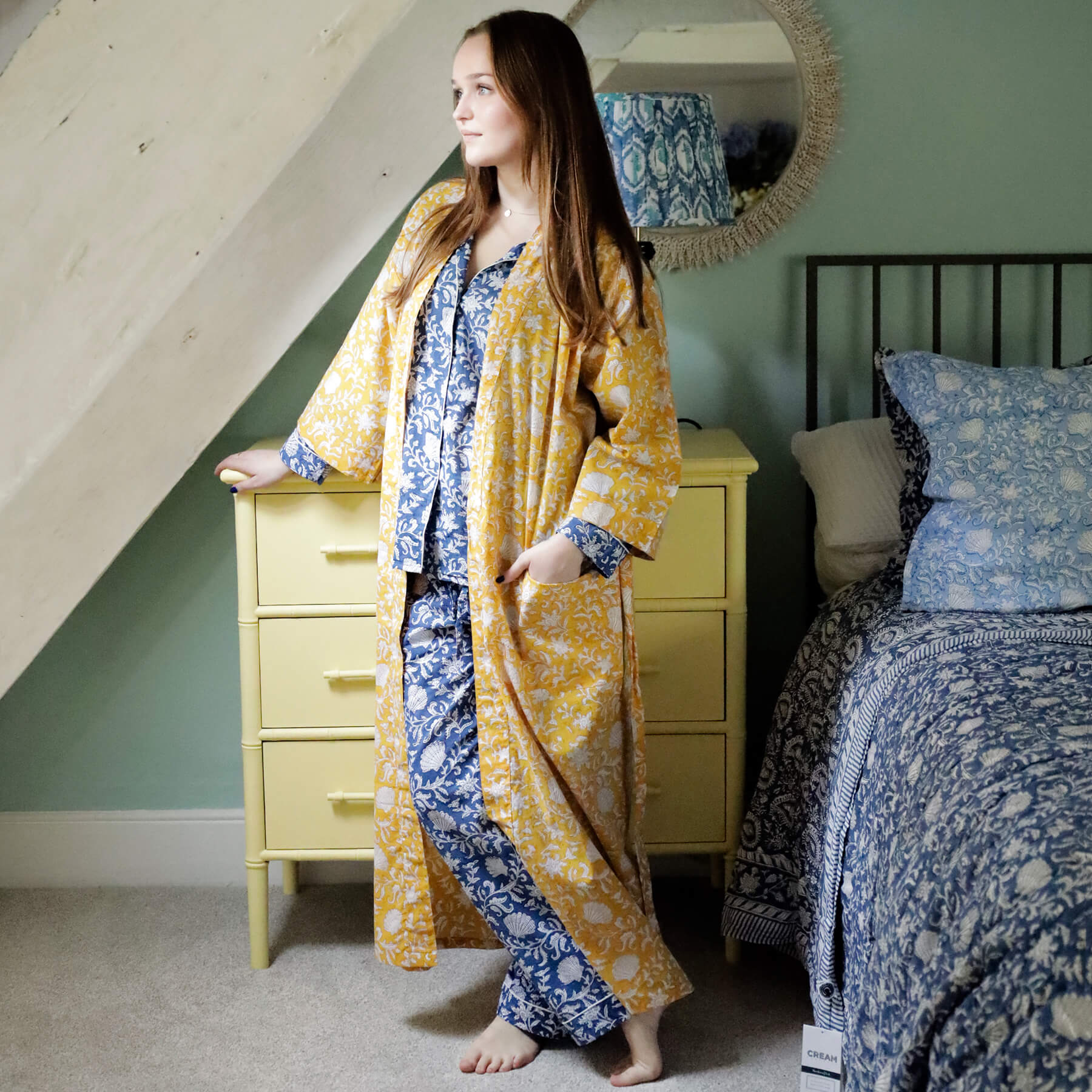 Young woman standing in a charming bedroom wearing a yellow block-printed dressing gown with white floral and seashell patterns over blue and white patterned pyjamas. The room features a yellow chest of drawers, a mirror, and a bed with matching blue block-printed bedding, creating a cozy and coordinated look