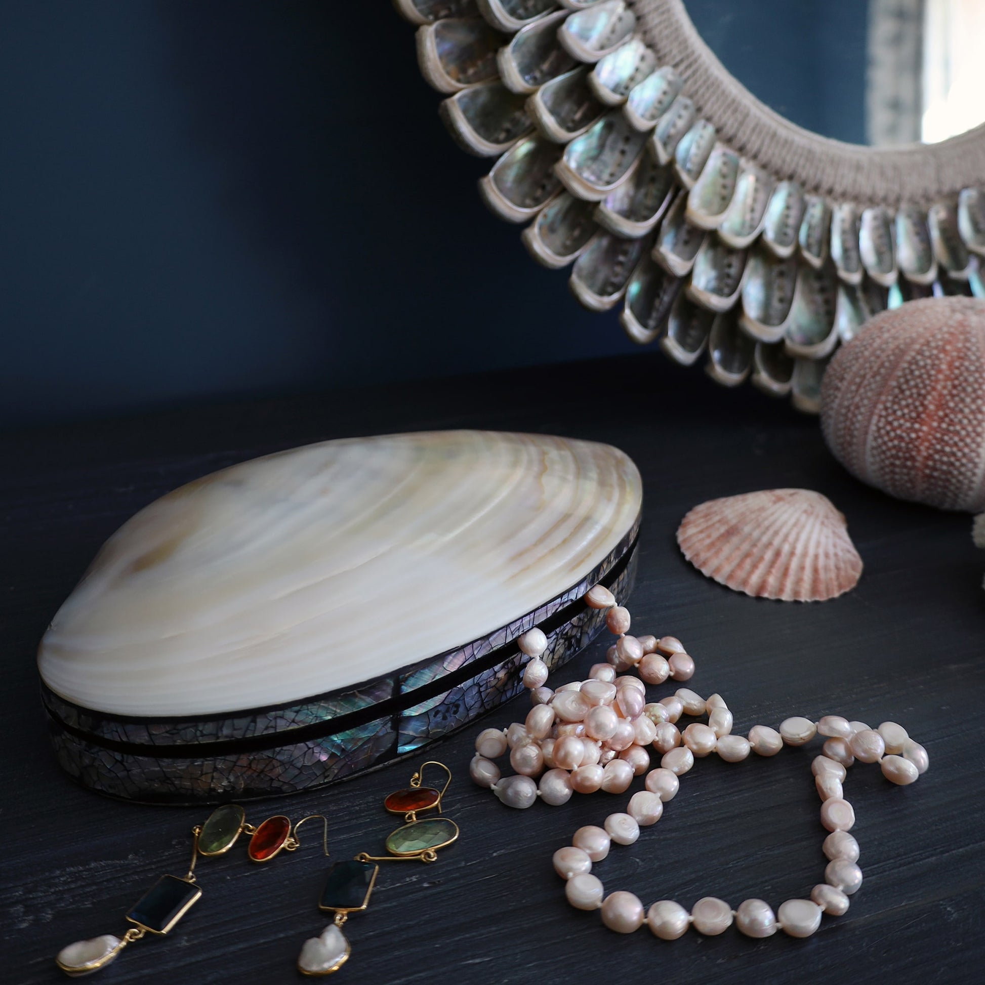 A close-up of a clam-shaped trinket box made from polished shell and edged with iridescent abalone inlay, resting on a dark wooden surface. A strand of pink freshwater pearls spills out of the box. Nearby are colorful gemstone drop earrings in gold settings, a textured sea urchin shell, a ridged clam shell, and part of a mirror with a decorative abalone shell frame.