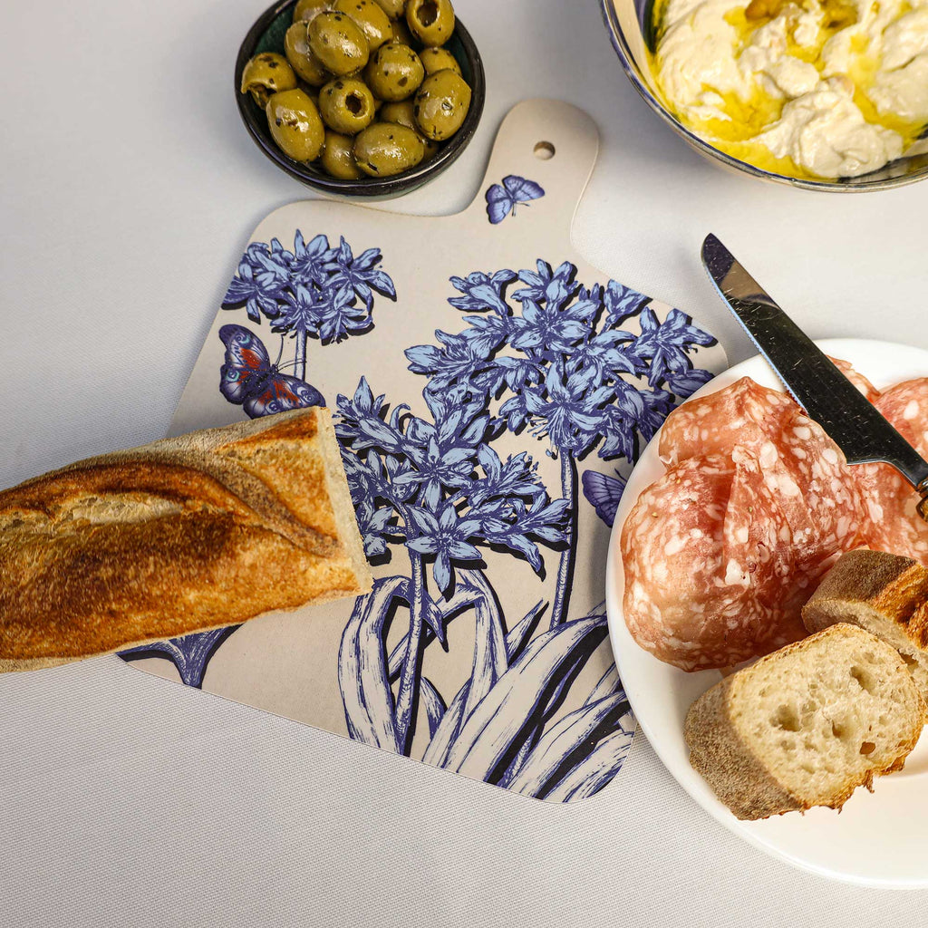 Cream Cornwall Agapanthus serving board featuring intricate blue botanical flowers and butterflies, styled with crusty bread, olives, cured meats and creamy dip on a white tablecloth.