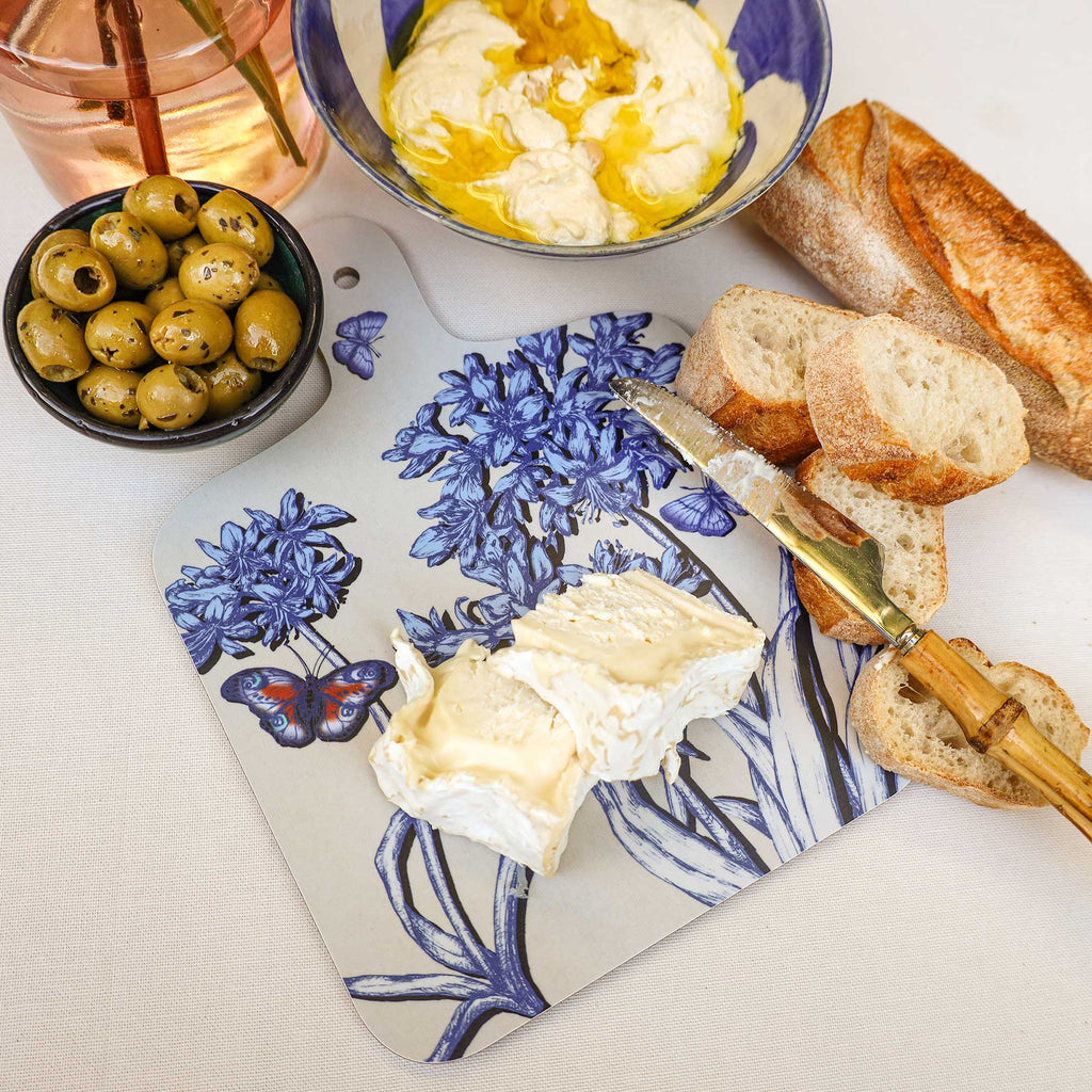 Cream Cornwall Agapanthus serving board featuring beautiful blue botanical flowers and butterflies, styled with creamy Brie cheese, crusty baguette, green olives, a cheese knife and a bowl of creamy dip on a white tablecloth.