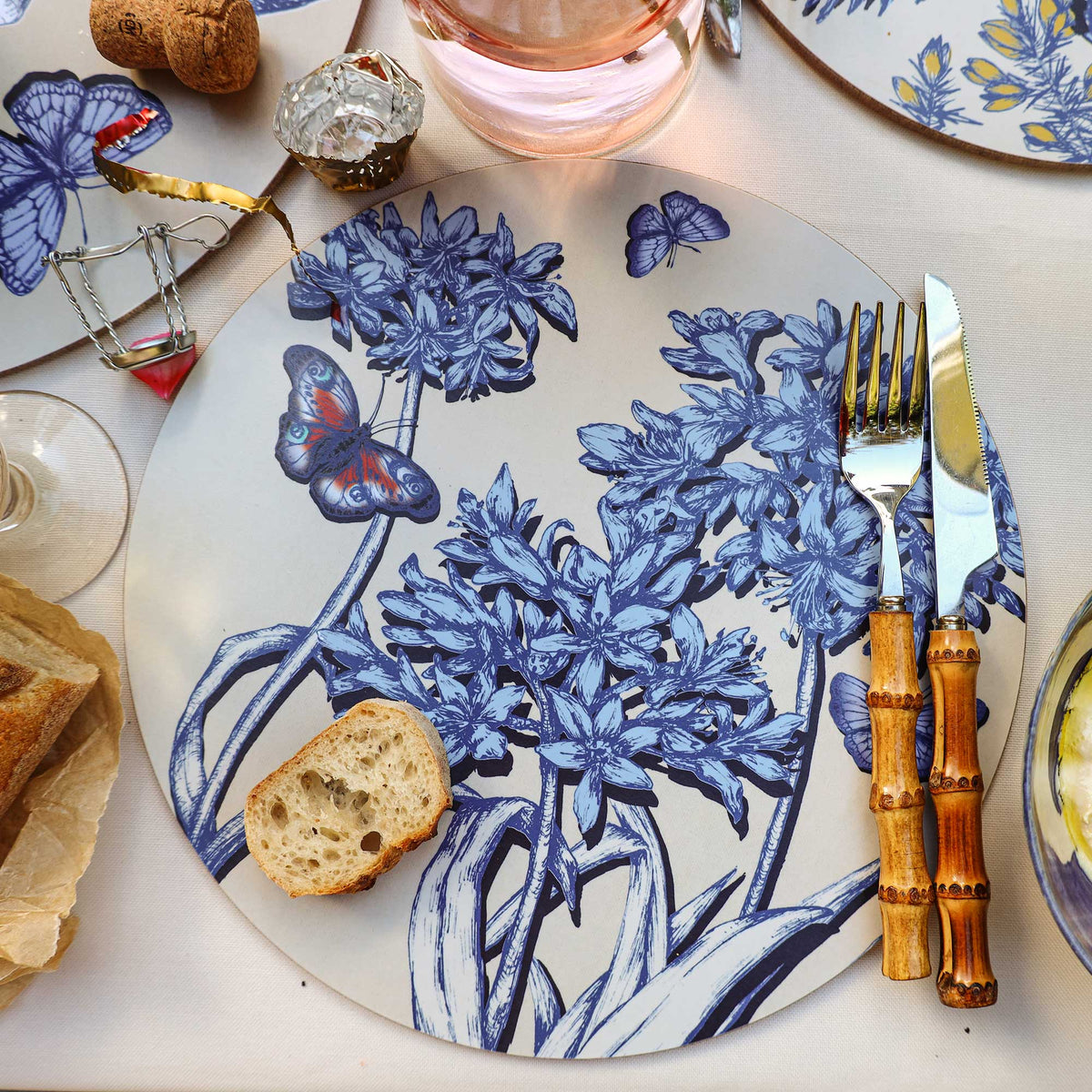 Overhead view of an elegant table setting featuring a round placemat with intricate blue botanical illustrations of clustered flowers, flowing leaves and delicate butterflies on a soft beige background. A colourful blue and orange butterfly adds a vibrant accent to the floral design. A slice of crusty bread rests on the placemat beside bamboo-handled cutlery, while a glass of rosé wine, a champagne cork and wire cage, and surrounding botanical tableware complete the arrangement on a cream linen tablecloth.