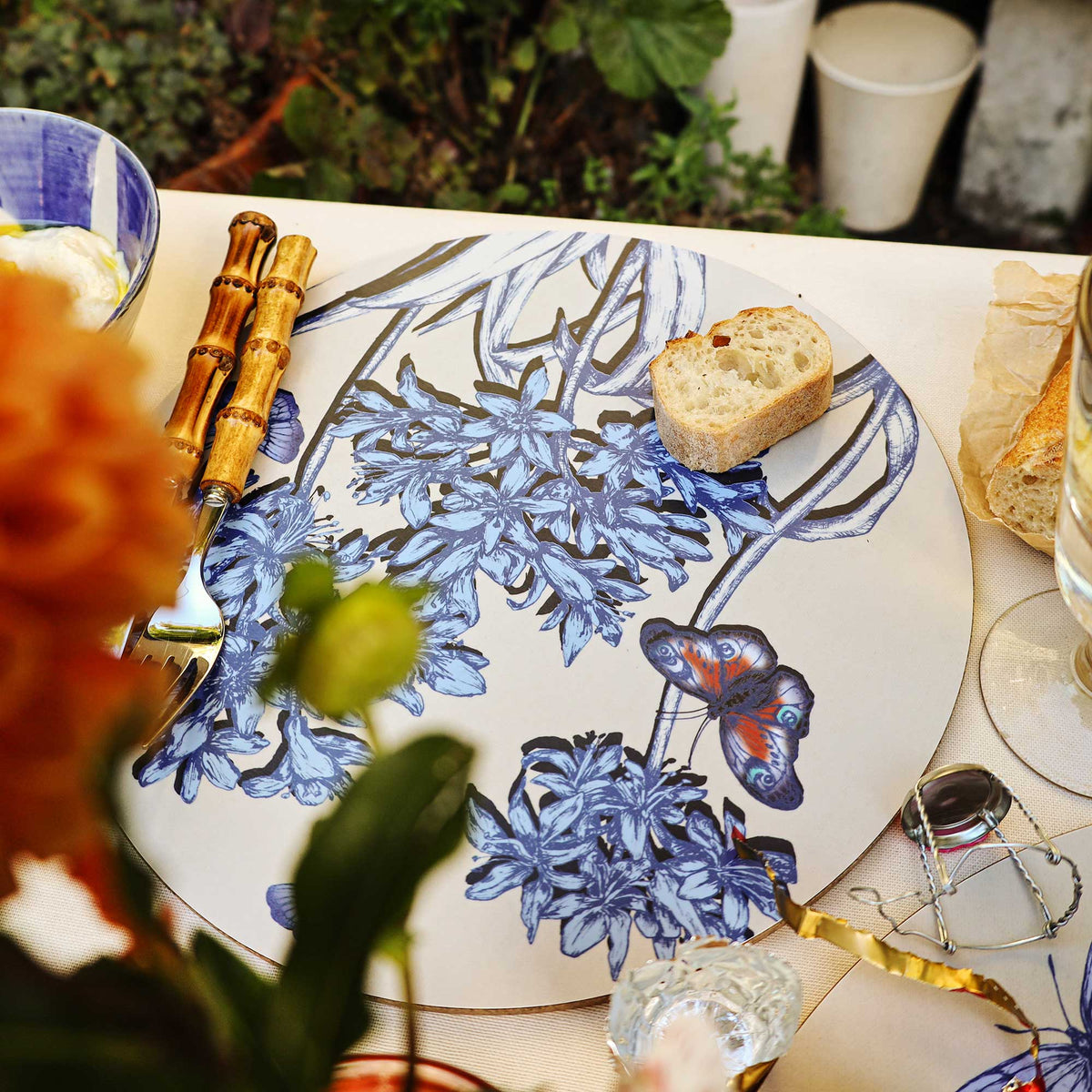 
Close-up of an elegant outdoor table setting featuring a round placemat with intricate blue botanical illustrations of clustered flowers, flowing stems and a colourful blue and orange butterfly on a cream background. A slice of crusty bread rests on the placemat beside bamboo-handled cutlery. A champagne cork and wire cage, glassware and additional bread surround the setting, while softly blurred orange flowers and green foliage frame the foreground.