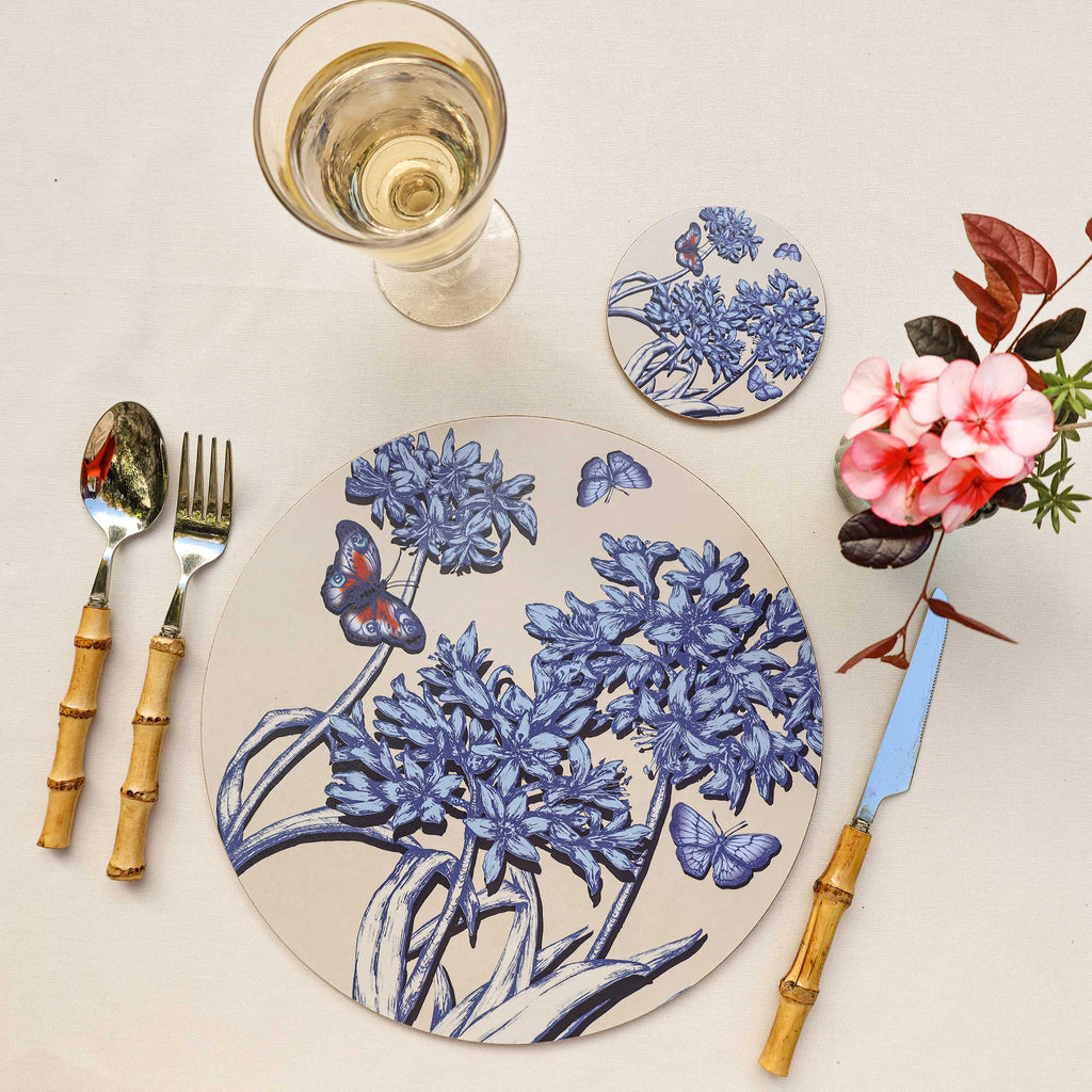 Overhead view of an elegant table setting featuring a round placemat with intricate blue botanical illustrations of flowering stems and butterflies, including a colourful blue and orange butterfly, on a soft beige background. A matching floral coaster sits above the placemat beside a glass of white wine. Bamboo-handled cutlery frames the arrangement, while delicate pink flowers and deep red foliage add a vibrant accent against the cream linen tablecloth.