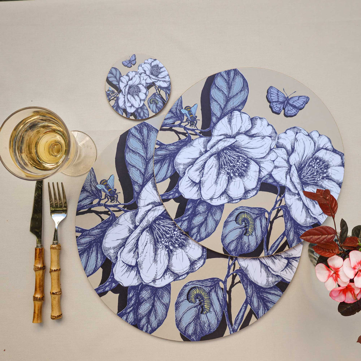 
Cream Cornwall round botanical placemats and matching coaster featuring elegant blue and white camellia flowers, butterflies, bees and detailed foliage on a soft cream background, styled with bamboo-handled cutlery, a glass of white wine and delicate pink flowers on a neutral linen tablecloth.