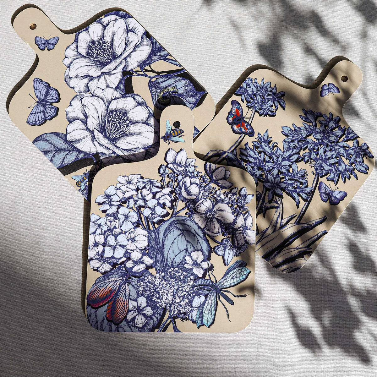 Three Cream Cornwall botanical serving boards featuring intricate blue and white illustrations of camellias, hydrangeas and agapanthus, with colourful butterflies, bees and dragonflies, arranged on a pale linen background with soft natural shadows.