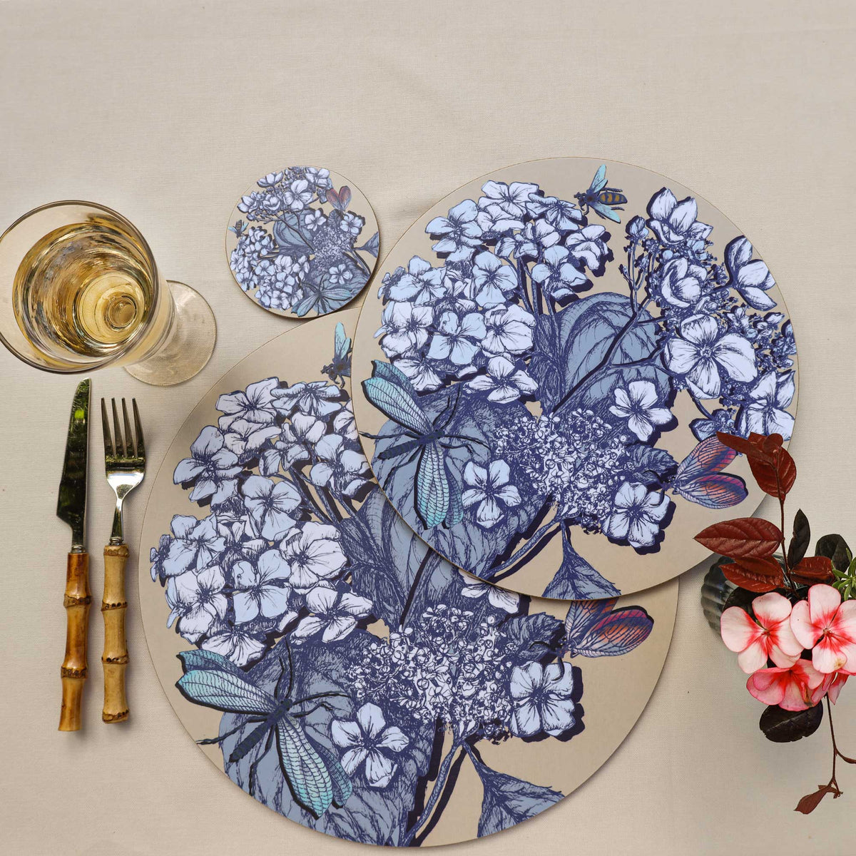 
Overhead view of two overlapping round placemats featuring intricate blue botanical illustrations of hydrangeas, leaves, dragonflies, bees and colourful butterflies on a soft beige background. A matching floral coaster, glass of white wine and bamboo-handled cutlery complement the arrangement, while delicate pink flowers and burgundy foliage add a pop of colour against the cream linen tablecloth.