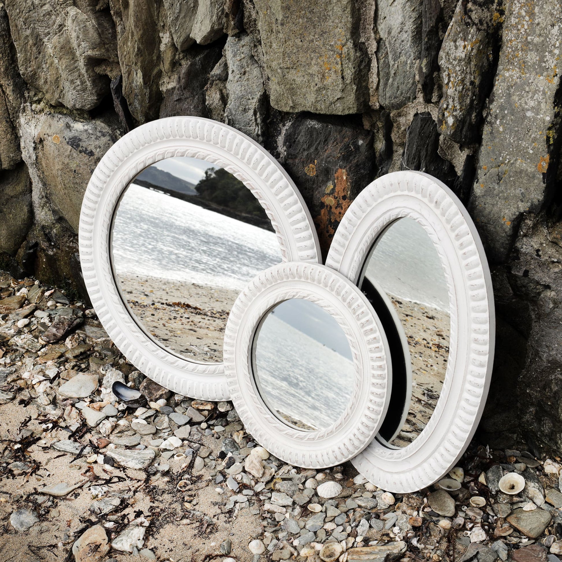3 whitewashed wooden mirrors with rope detail carving propped up against the harbour wall on the beach with the shoreline reflected in it.