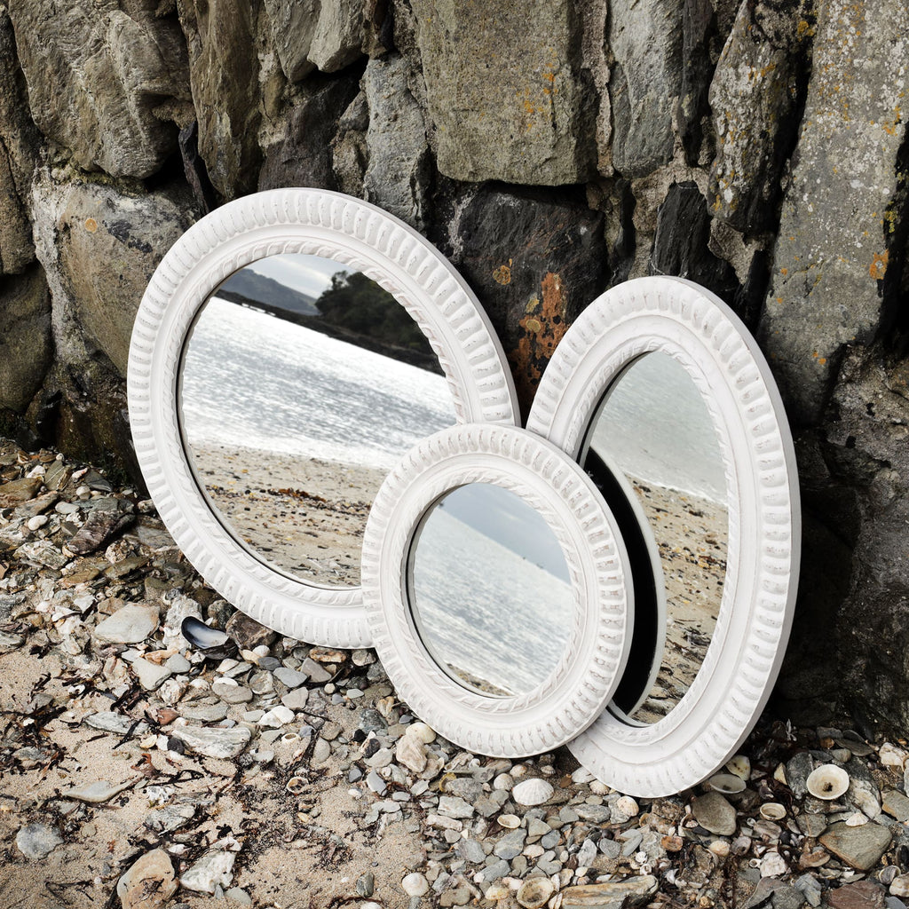3 whitewashed wooden mirrors with rope detail carving propped up against the harbour wall on the beach with the shoreline reflected in it.