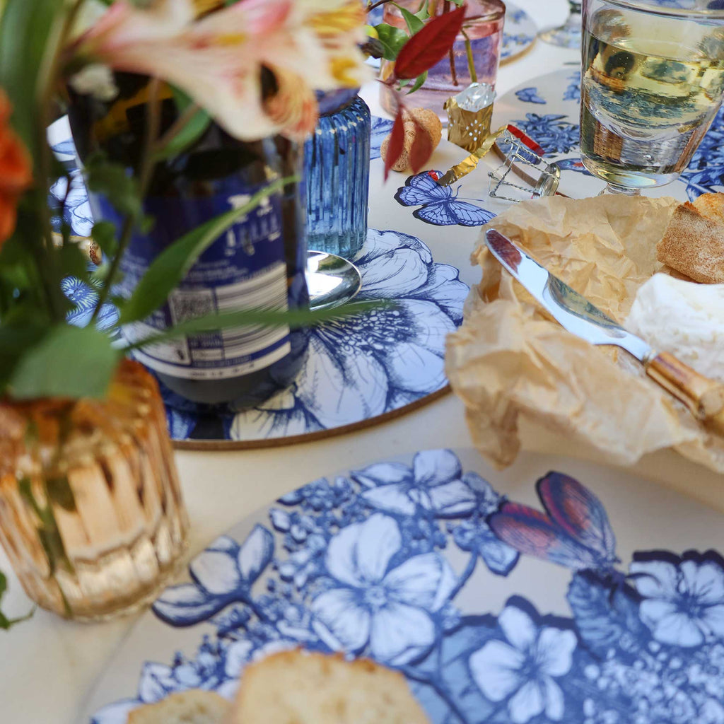 
Close-up of an elegant dining tablescape featuring round placemats with intricate blue botanical illustrations of flowers, leaves and butterflies. A wine bottle, glass of white wine, sliced bread and soft cheese on crinkled parchment paper are arranged alongside bamboo-handled cutlery. Delicate pink and orange flowers, lush greenery and decorative glassware complete the setting on a cream tablecloth.