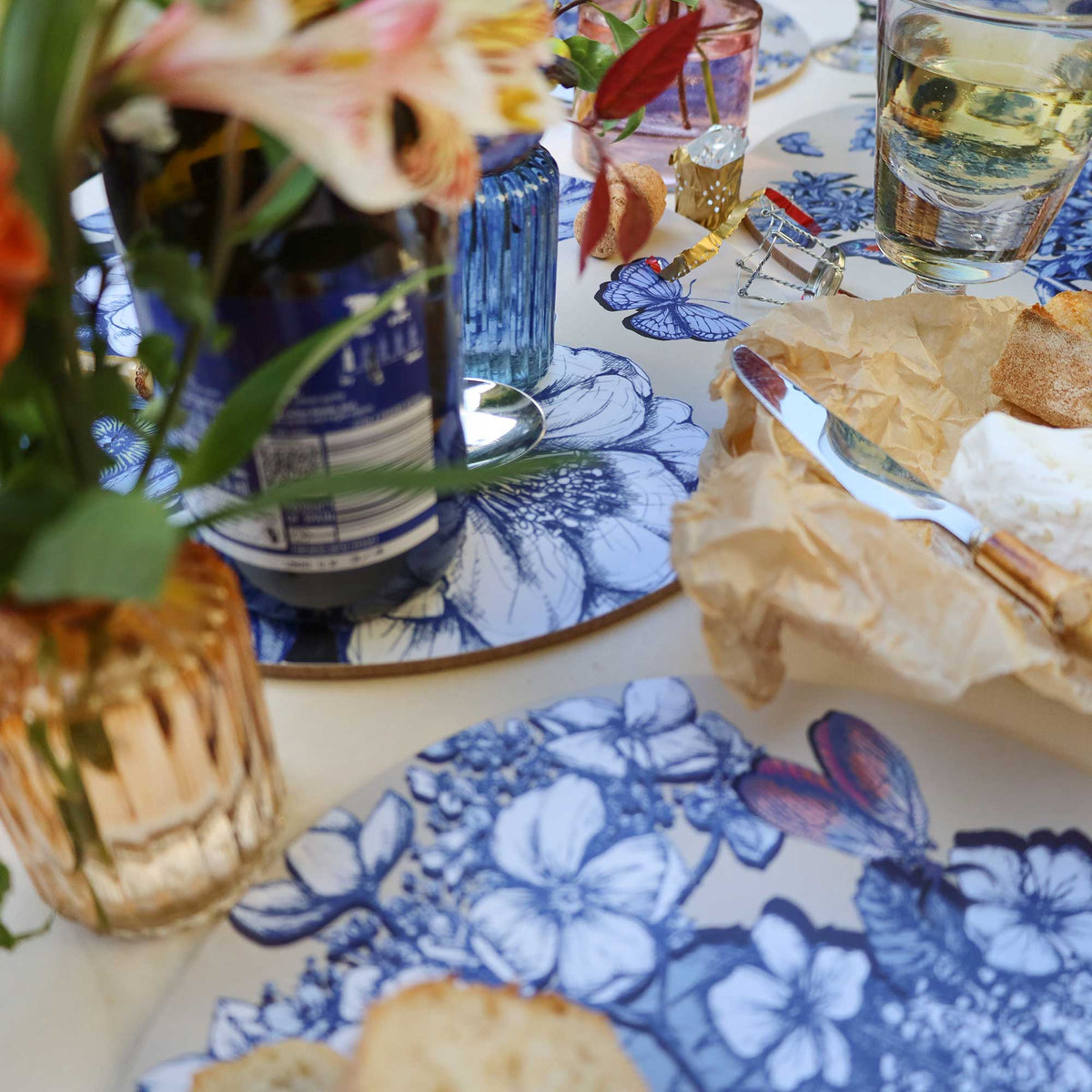 
Close-up of an elegant dining tablescape featuring round placemats with intricate blue botanical illustrations of flowers, leaves and butterflies. A wine bottle, glass of white wine, sliced bread and soft cheese on crinkled parchment paper are arranged alongside bamboo-handled cutlery. Delicate pink and orange flowers, lush greenery and decorative glassware complete the setting on a cream tablecloth.
