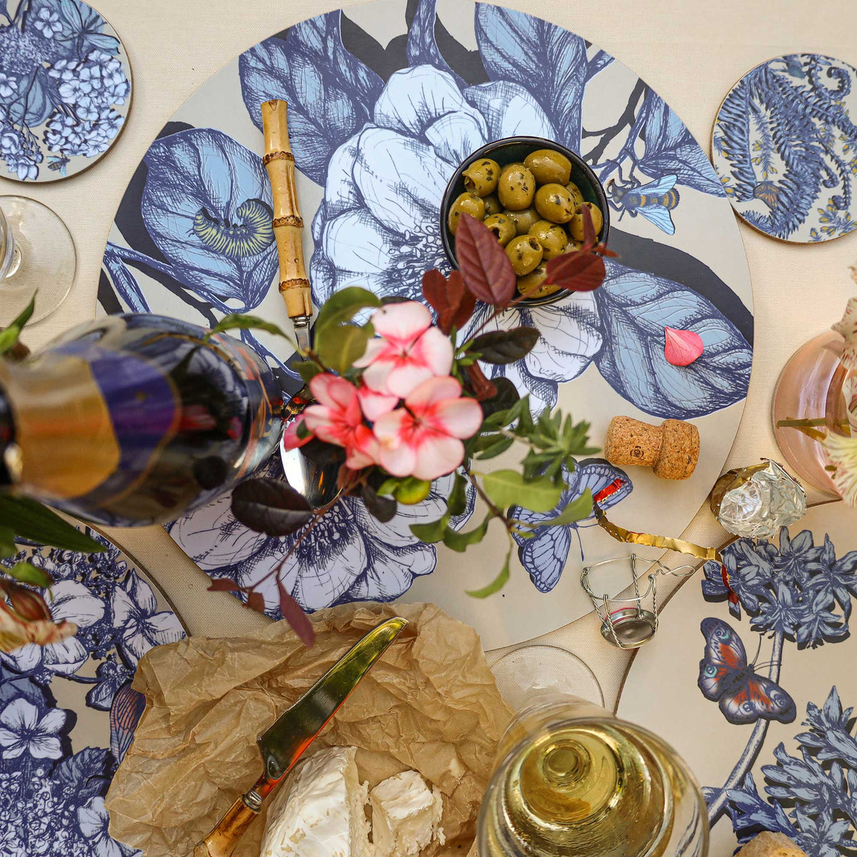 
Overhead view of an elegant Cream Cornwall table setting featuring round placemats and matching coasters decorated with intricate blue botanical illustrations, butterflies and insects on a cream background. Styled with soft cheese on brown parchment, a bamboo-handled cheese knife, green olives, a glass of white wine, pink flowers and decorative table accessories on a neutral linen tablecloth.