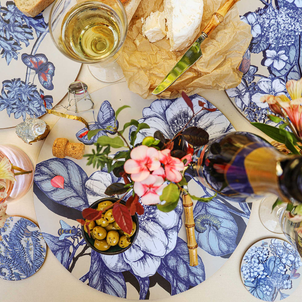 Overhead view of an elegant Cream Cornwall table setting featuring round placemats and matching coasters decorated with intricate blue botanical illustrations, butterflies and insects on a cream background. Styled with soft cheese on brown parchment, a bamboo-handled cheese knife, green olives, a glass of white wine, pink flowers and decorative table accessories on a neutral linen tablecloth.
