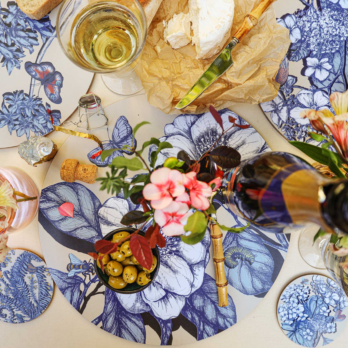 Overhead view of an elegant Cream Cornwall table setting featuring round placemats and matching coasters decorated with intricate blue botanical illustrations, butterflies and insects on a cream background. Styled with soft cheese on brown parchment, a bamboo-handled cheese knife, green olives, a glass of white wine, pink flowers and decorative table accessories on a neutral linen tablecloth.