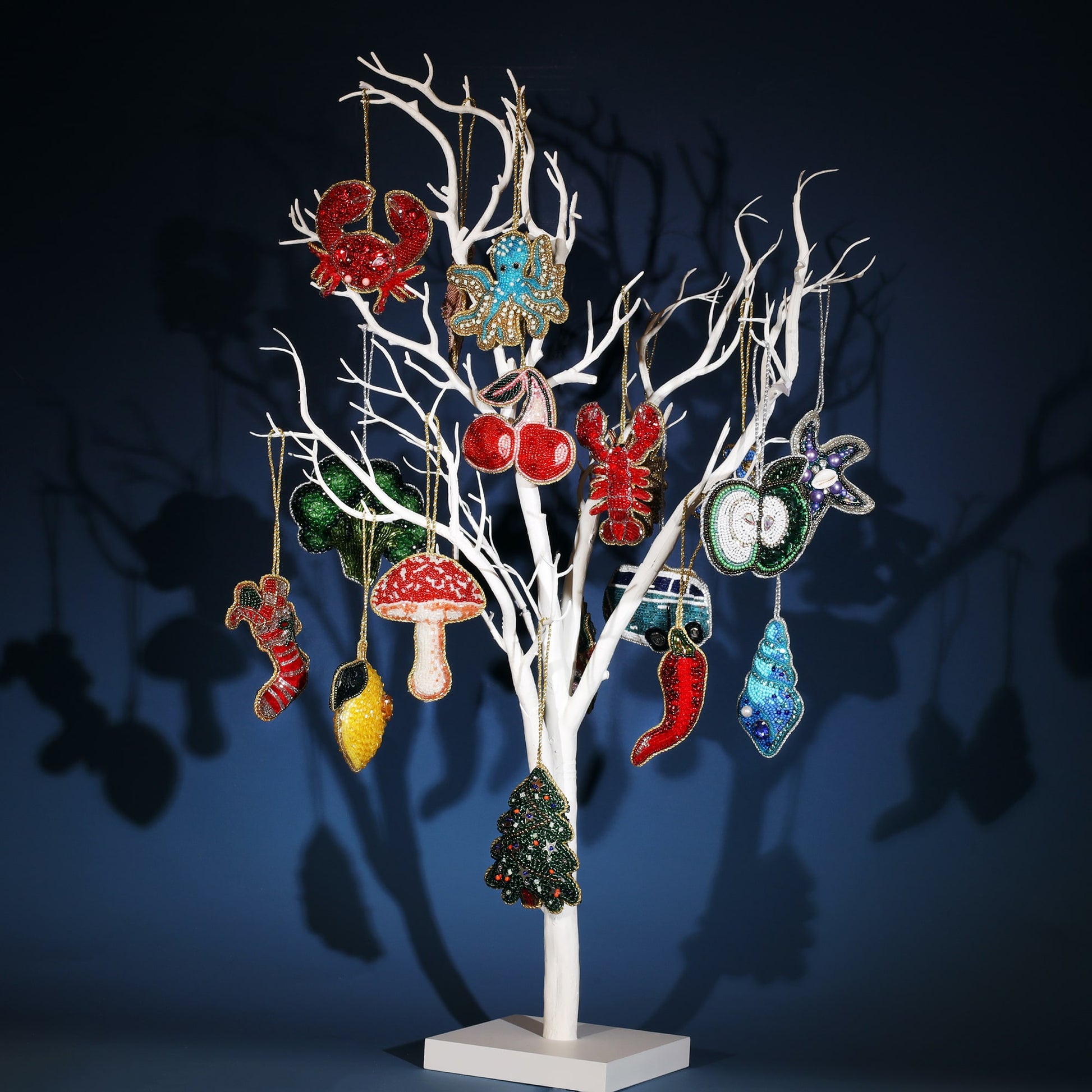  collection of vibrant beaded Christmas ornaments hanging on a white decorative tree. The ornaments include various designs such as a red lobster, blue octopus, mushroom, cherries, chili pepper, green apple, and a Christmas tree. Each ornament is intricately detailed with colorful beads and sparkling embellishments. The display is set against a dark blue background, casting playful shadows of the ornaments and tree.